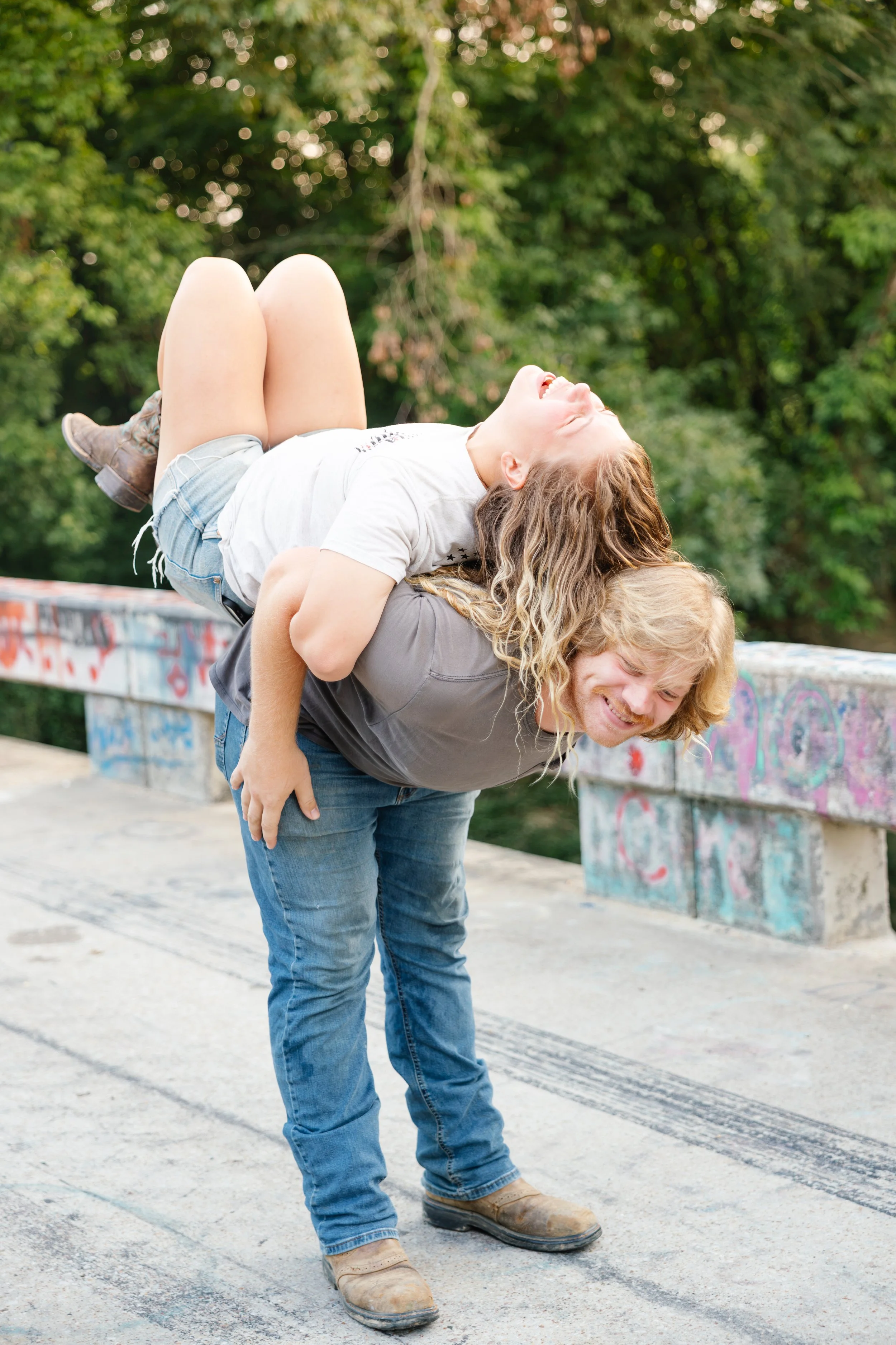 A man and a woman are playing on a skatepark ledge, with the woman lying across the man's shoulders and laughing. The man dressed in jeans and boots is bending forward in a playful pose, while the woman in casual clothing is on his back. The background features graffiti-covered concrete and green trees.