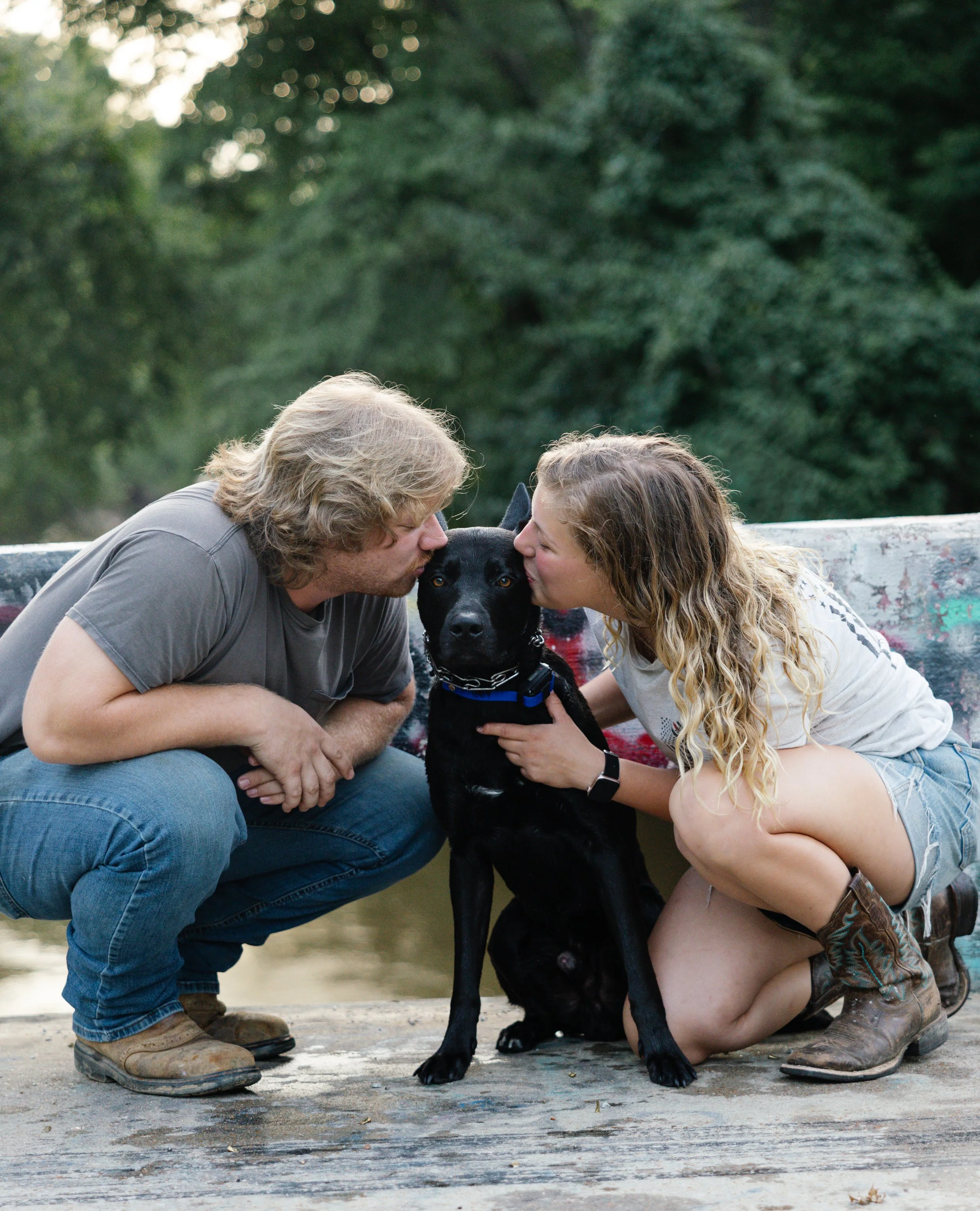 A man and woman are kissing a black dog on the head while squatting on the ground outdoors. The woman is wearing a white shirt, and the man is wearing a gray shirt and jeans. The background is blurred with green trees.
