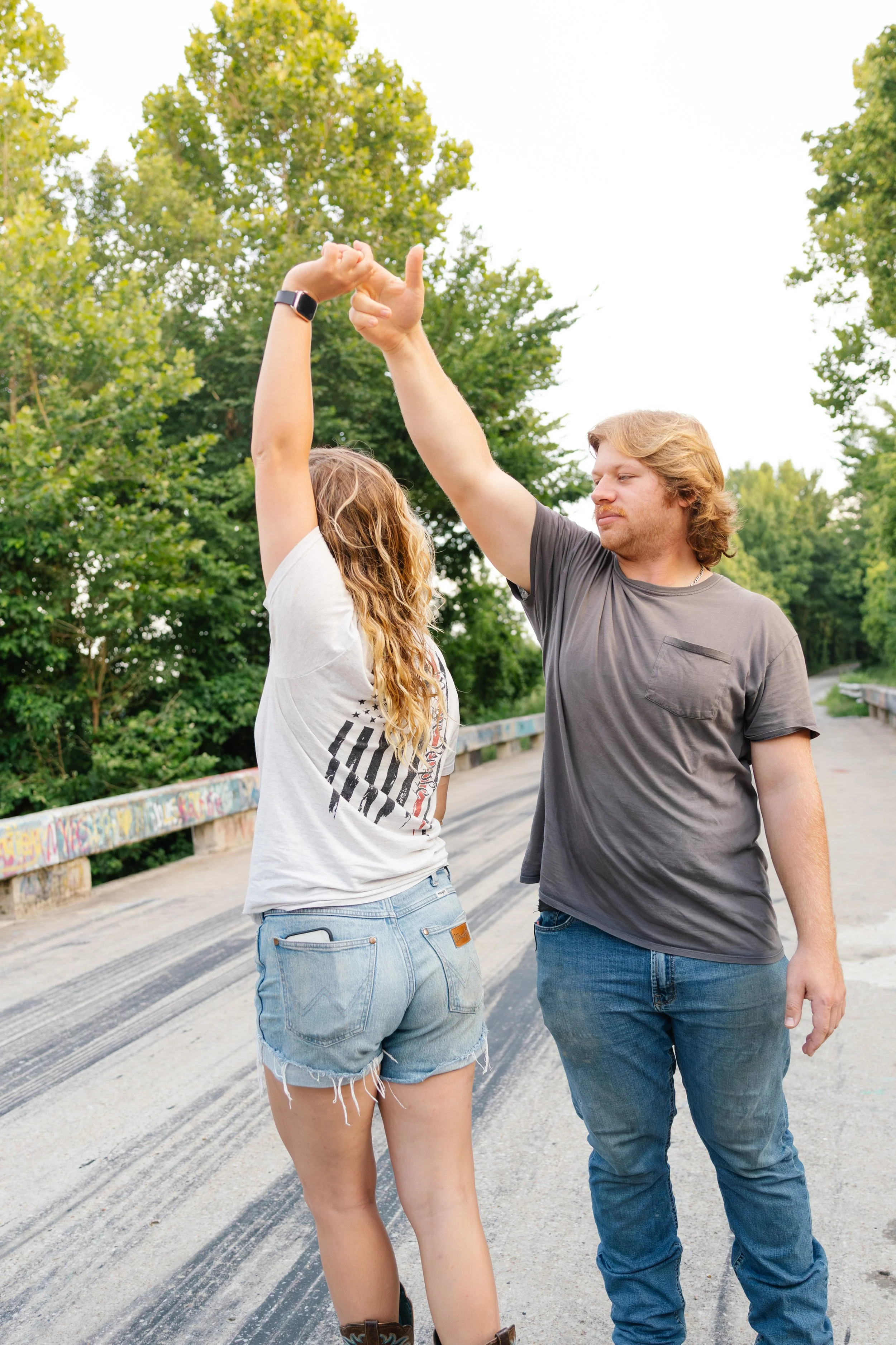 A man and a woman are dancing or celebrating together outdoors on a bridge with graffiti, surrounded by green trees.