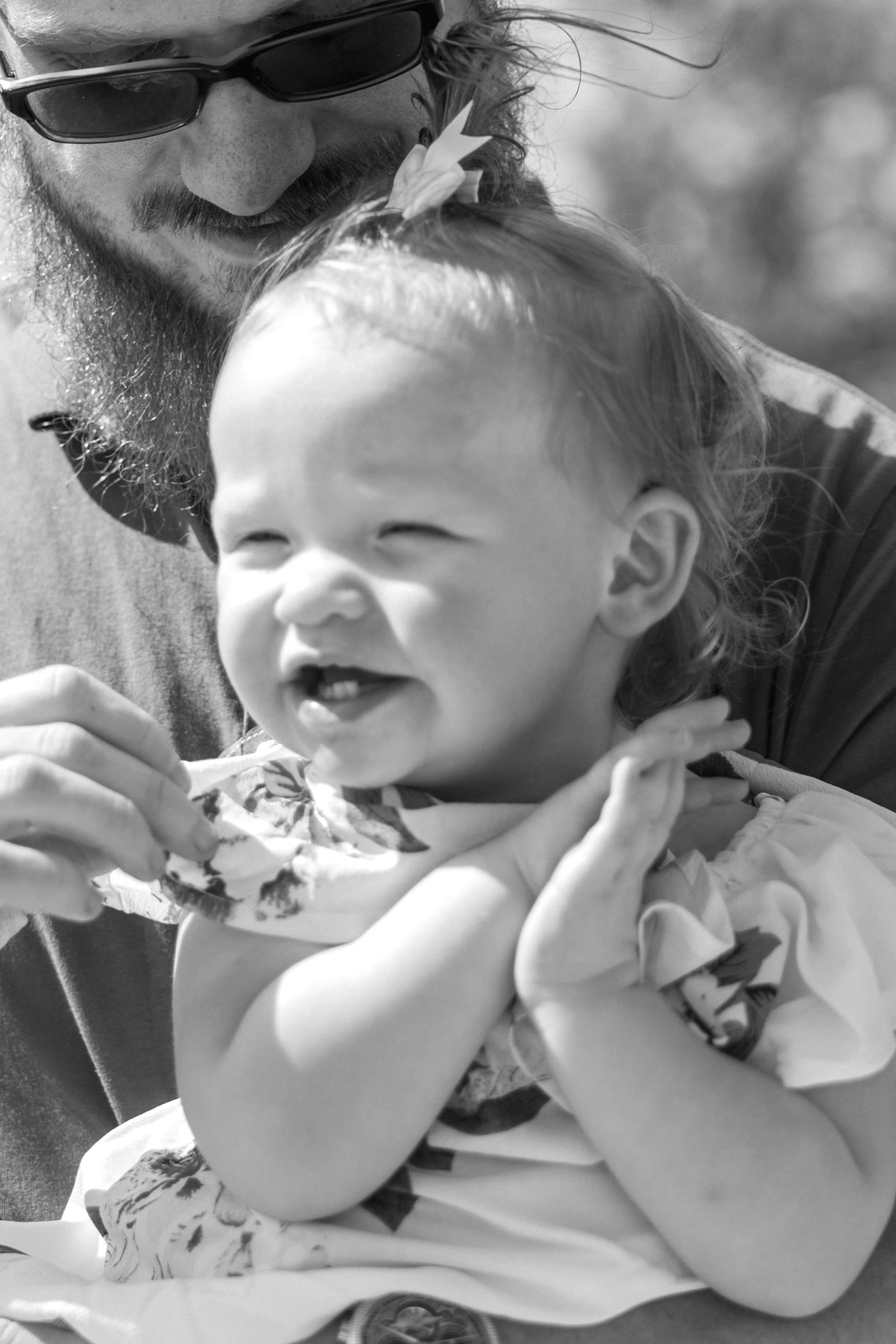 A man with sunglasses, a beard, and mustache kissing a small laughing girl with light curly hair and light skin, who is wearing a floral dress and clasping her hands together. The background is blurred.