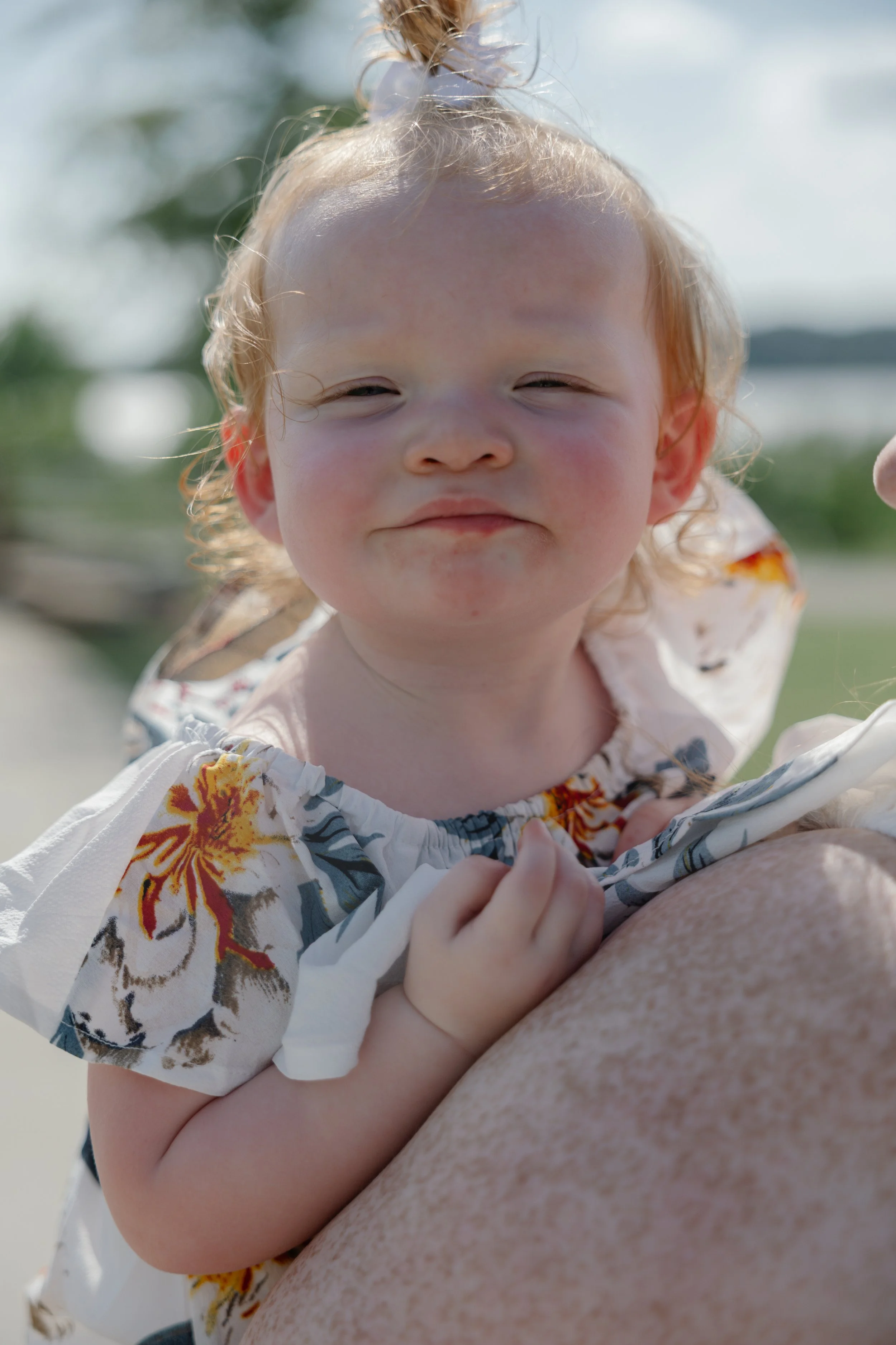 A young child with red hair and a floral dress smiling while being held outdoors on a sunny day.