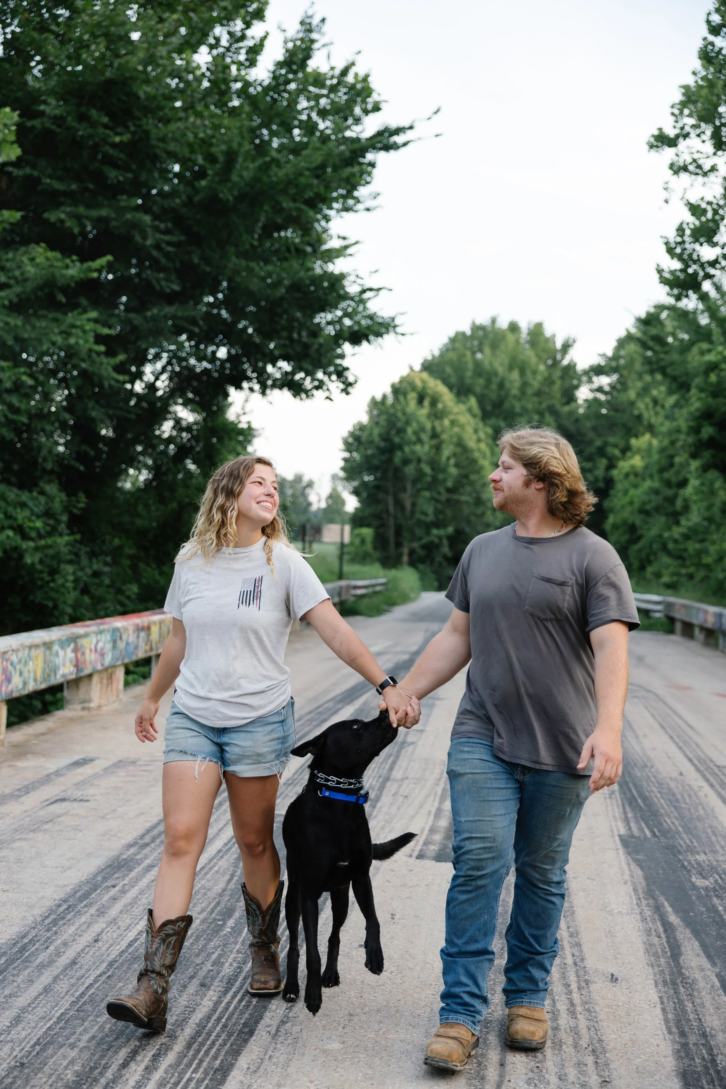 A young woman and man are walking on a bridge holding hands with a black dog between them. The woman is smiling and looking at the man. The woman is wearing a gray t-shirt, jean shorts, and cowboy boots. The man is wearing a gray t-shirt, jeans, and 