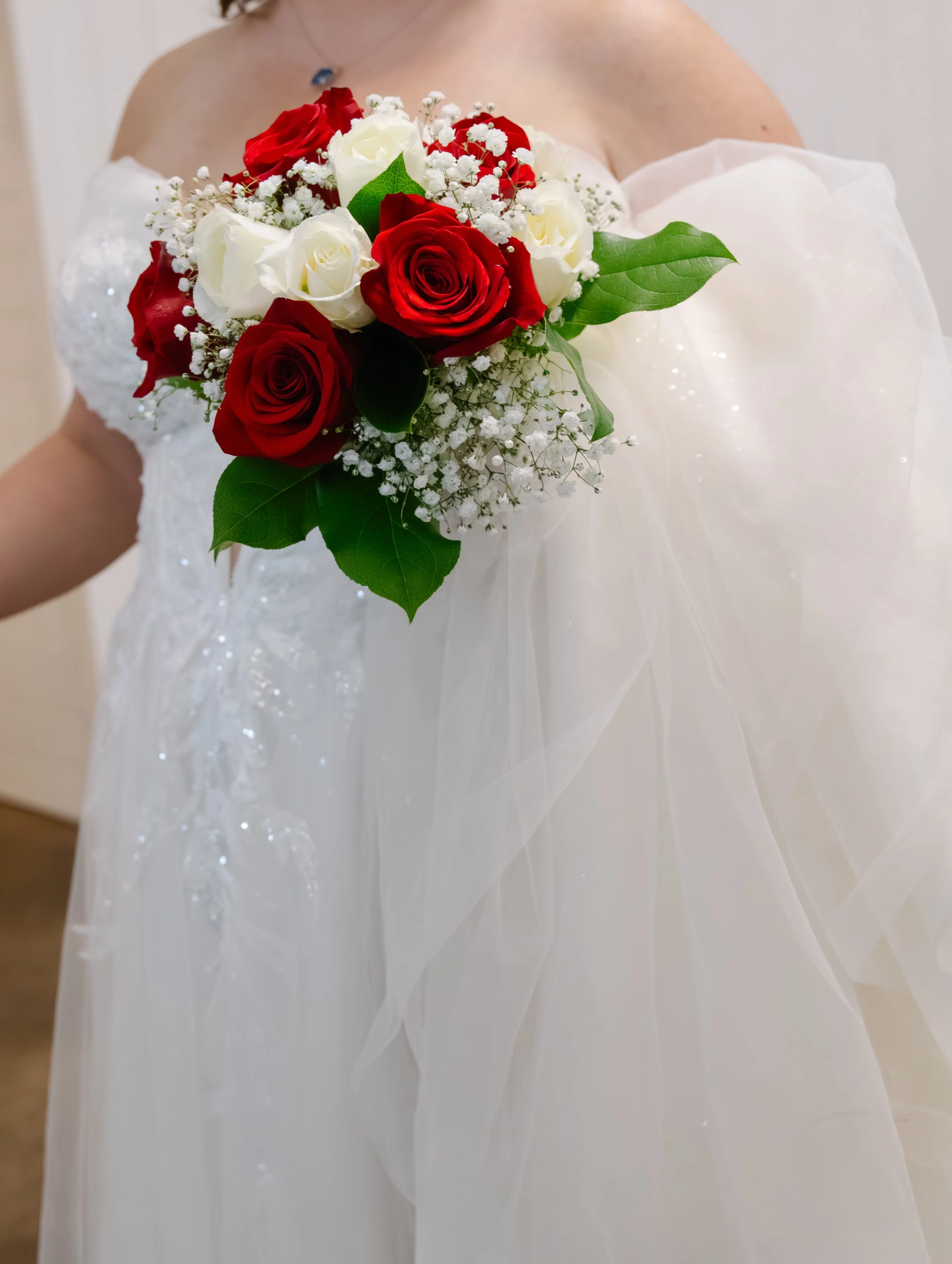 Close-up of a bouquet of red and white roses with baby's breath and large green leaves, held by a woman dressed in a white, off-shoulder wedding gown.