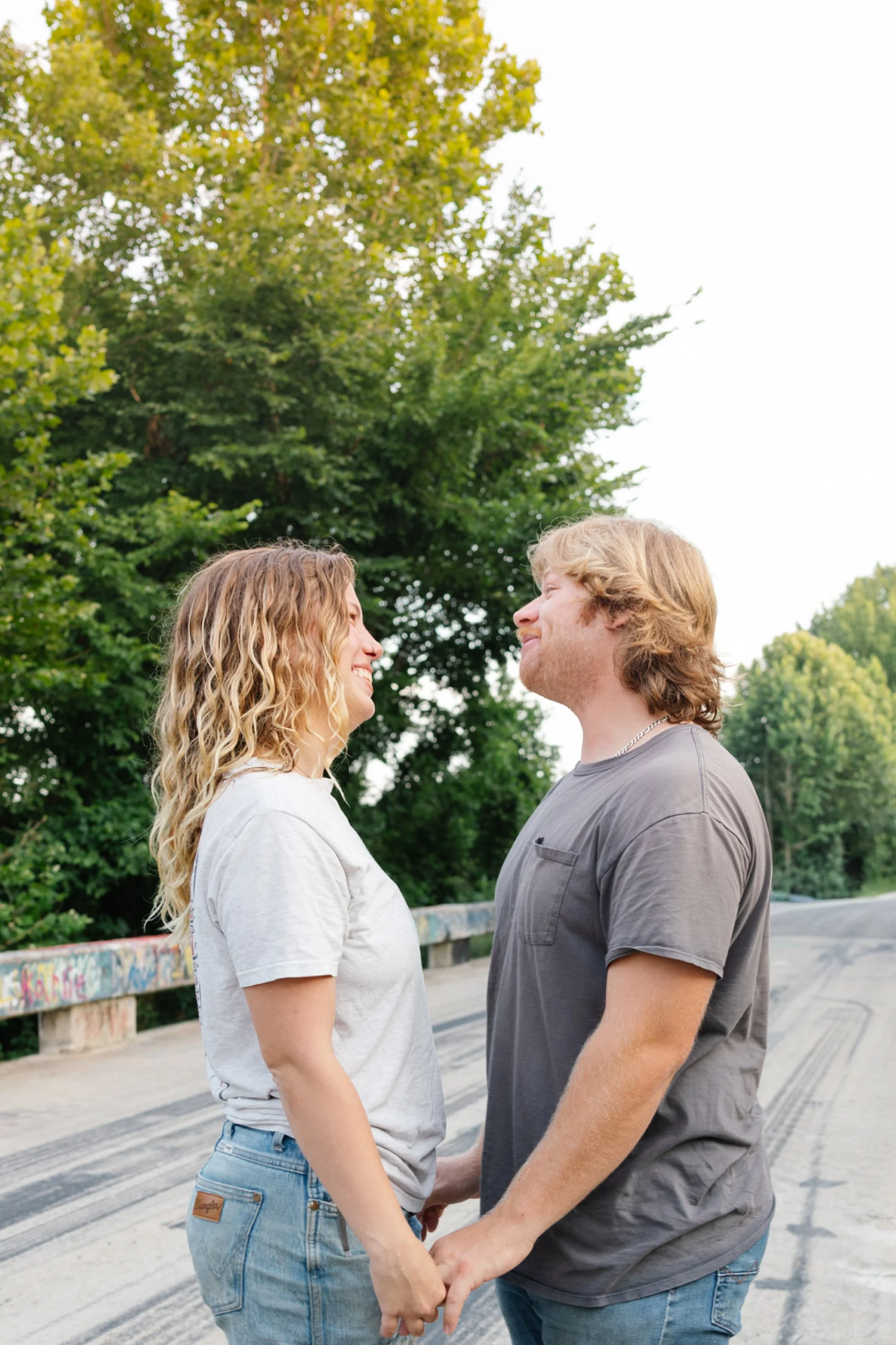 A couple stands face to face holding hands while smiling at each other outdoors on a bridge with graffiti. The woman has curly blonde hair, wears a white t-shirt and jeans, and the man has wavy blonde hair, wears a gray t-shirt and jeans. Green trees