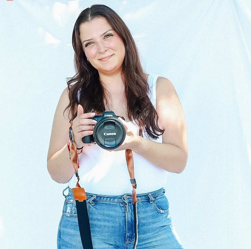 A young woman with long dark hair taking a selfie with a Canon camera, smiling slightly, against a bright, possibly outdoor, background.