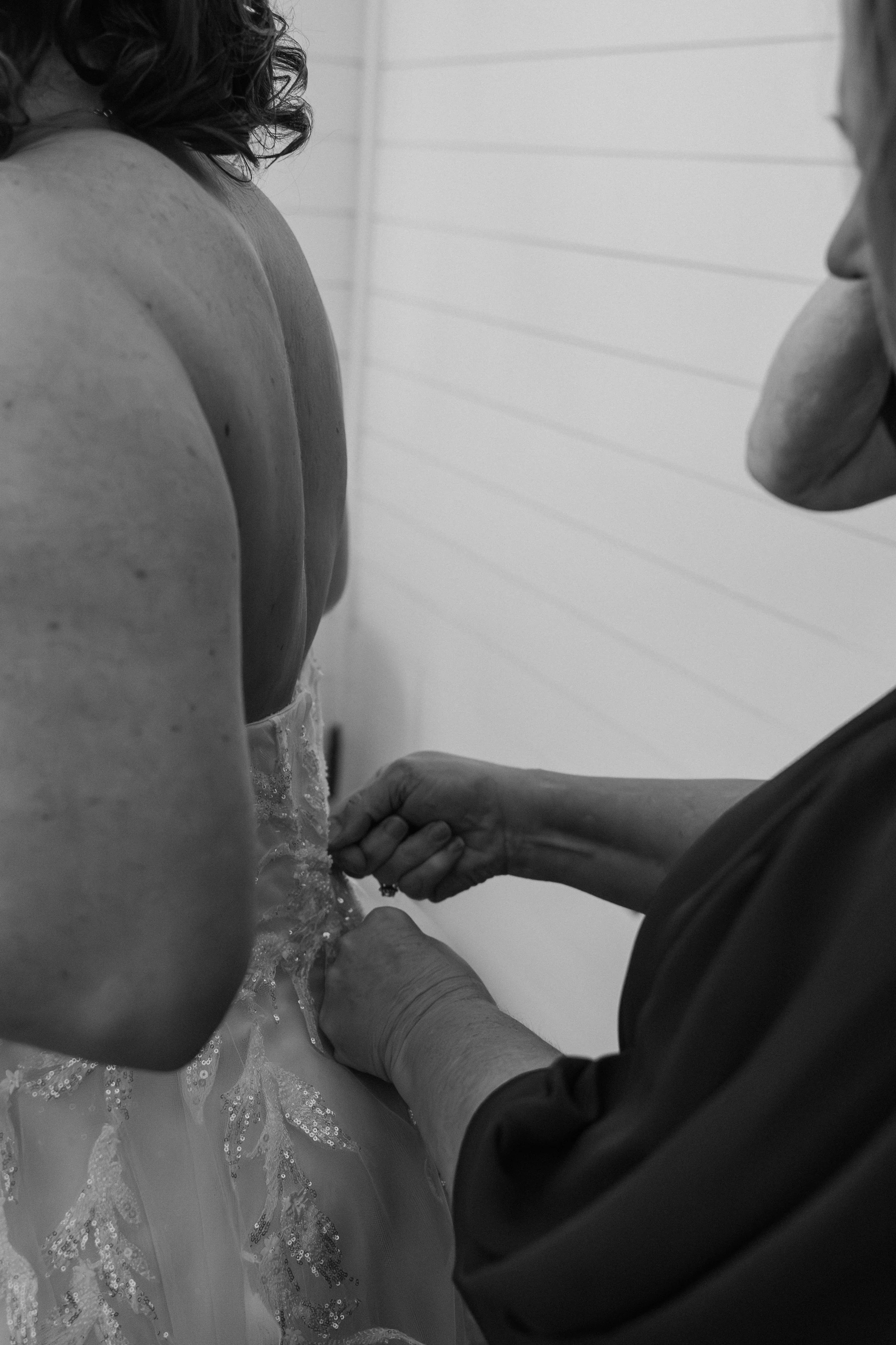 A close-up black-and-white photo of a person with long, curly hair, wearing a dress with lace and sequins, being adjusted by a second person with glasses and short hair. The second person's hands are fastening or fixing something on the first person'
