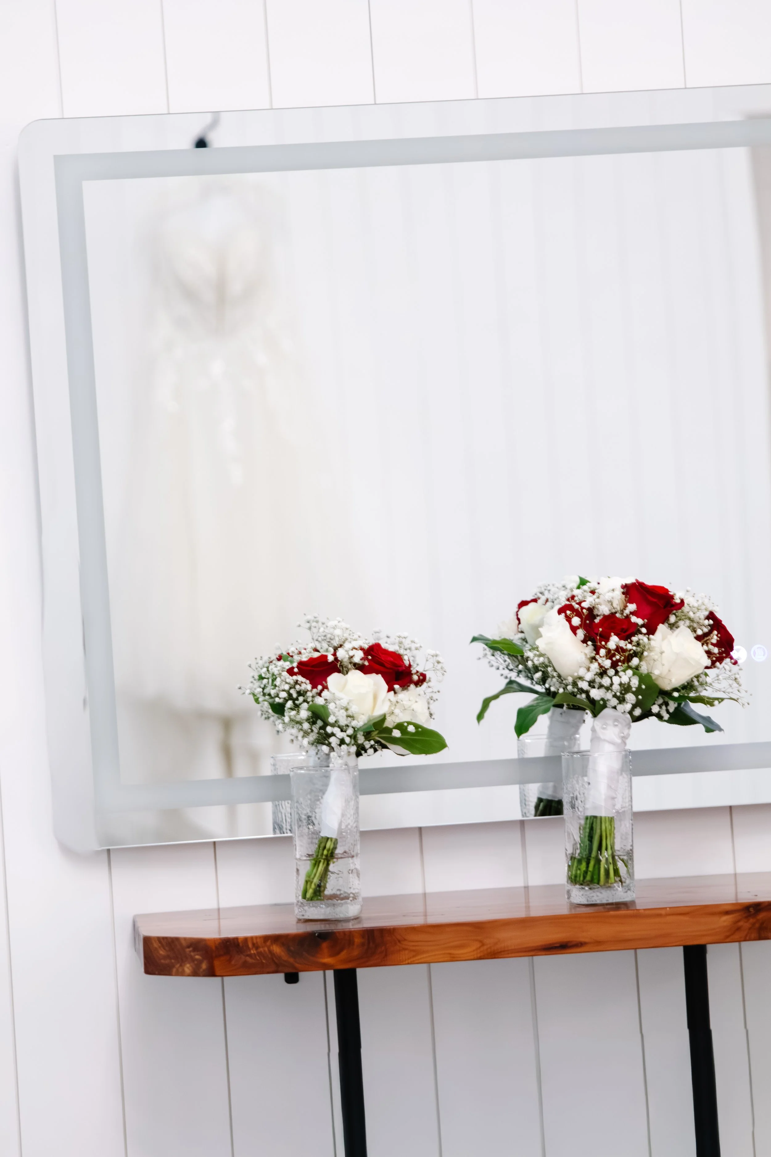 Three glass vases filled with white roses, red roses, baby's breath, and green leaves, positioned on a wooden table in front of an oval mirror with a white frame in a white room.