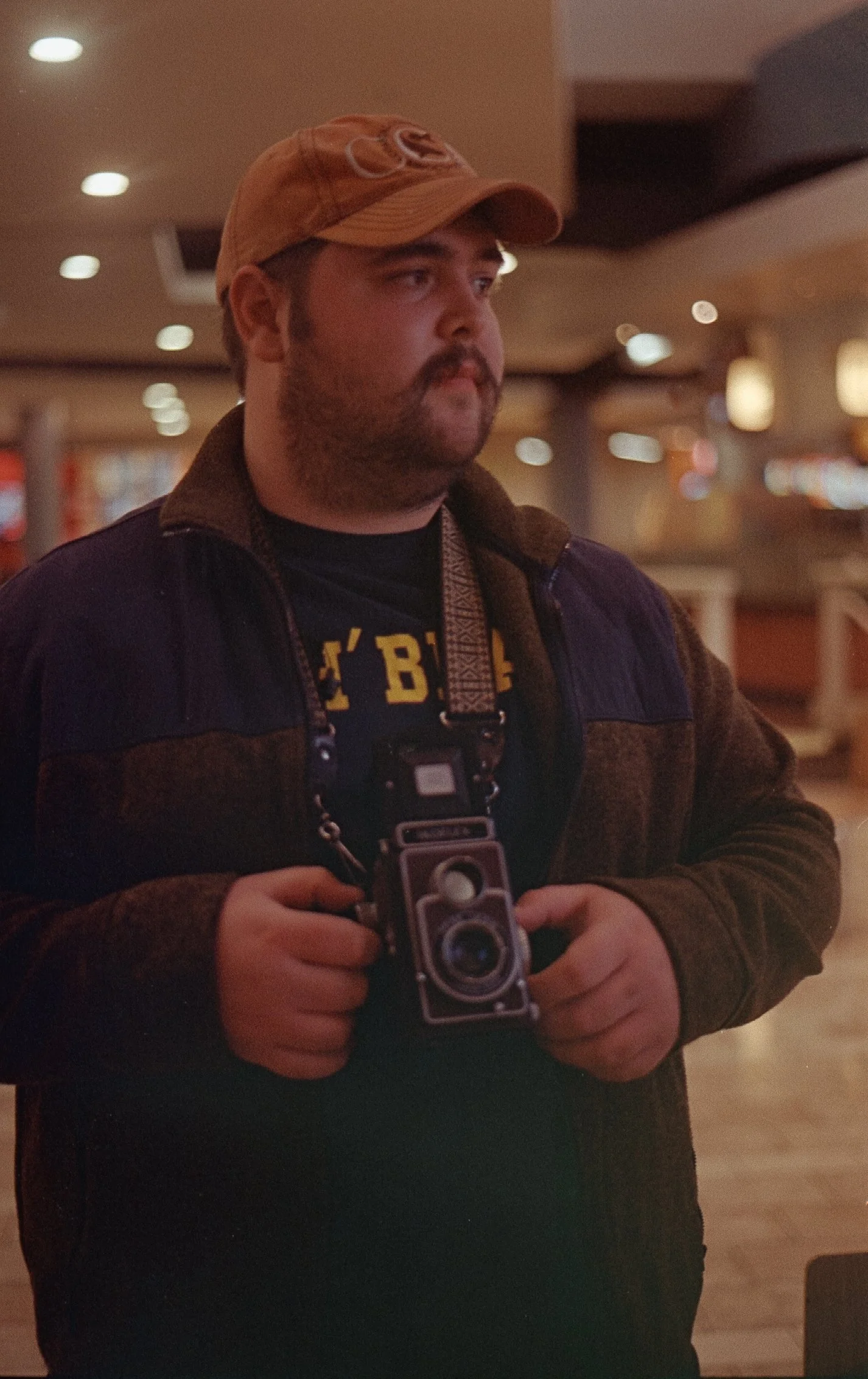 A man wearing a tan cap and dark jacket holding a vintage camera indoors with blurred lights and background.