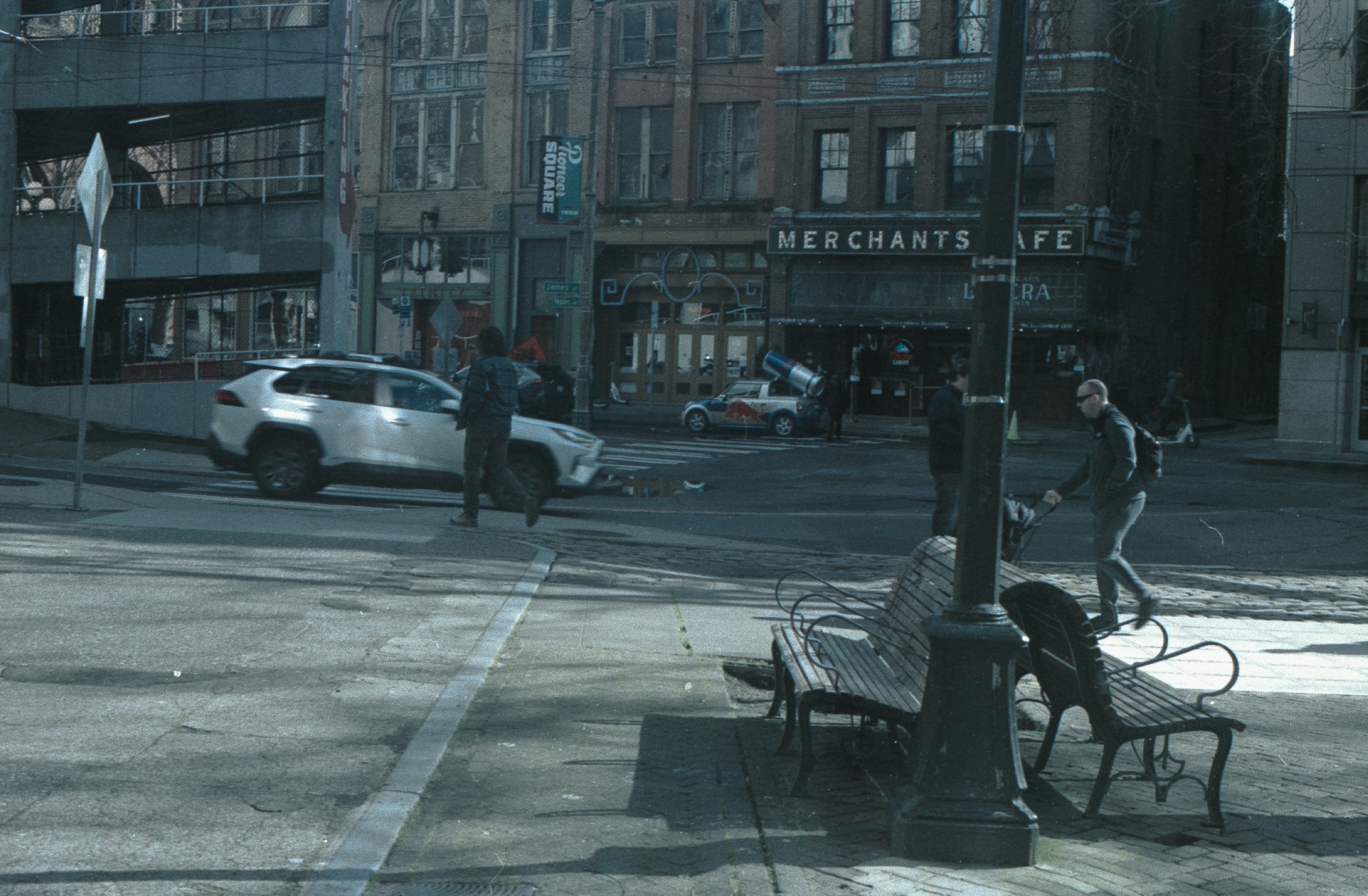 City street scene with pedestrians, parked cars, and a building with a sign reading 'MERCHANTS CAFE'.