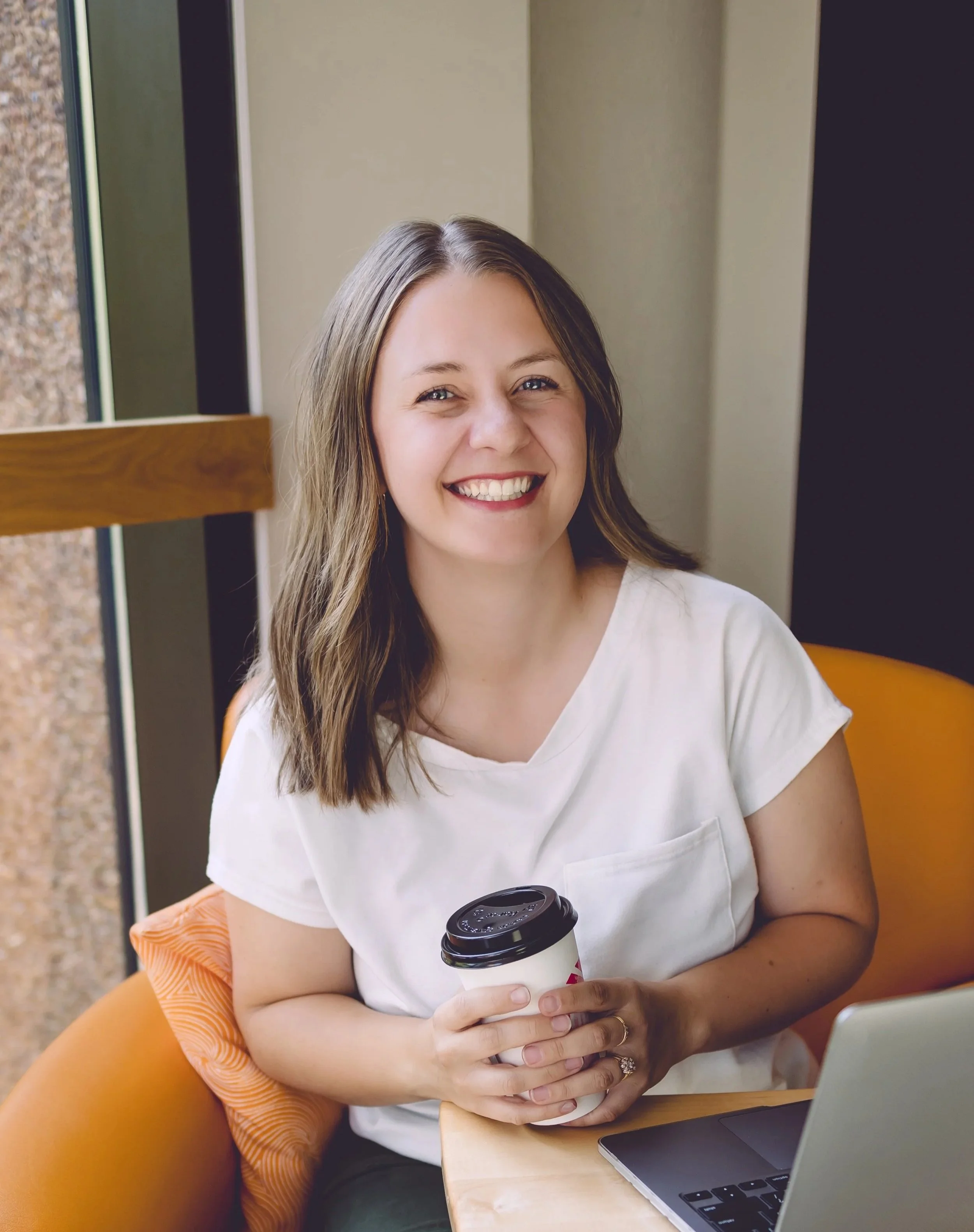 Kassi Strong, founder of Wellhouse marketing in Billings, Montana sitting at a table, holding a coffee cup, in front of a laptop, in a cozy indoor setting.