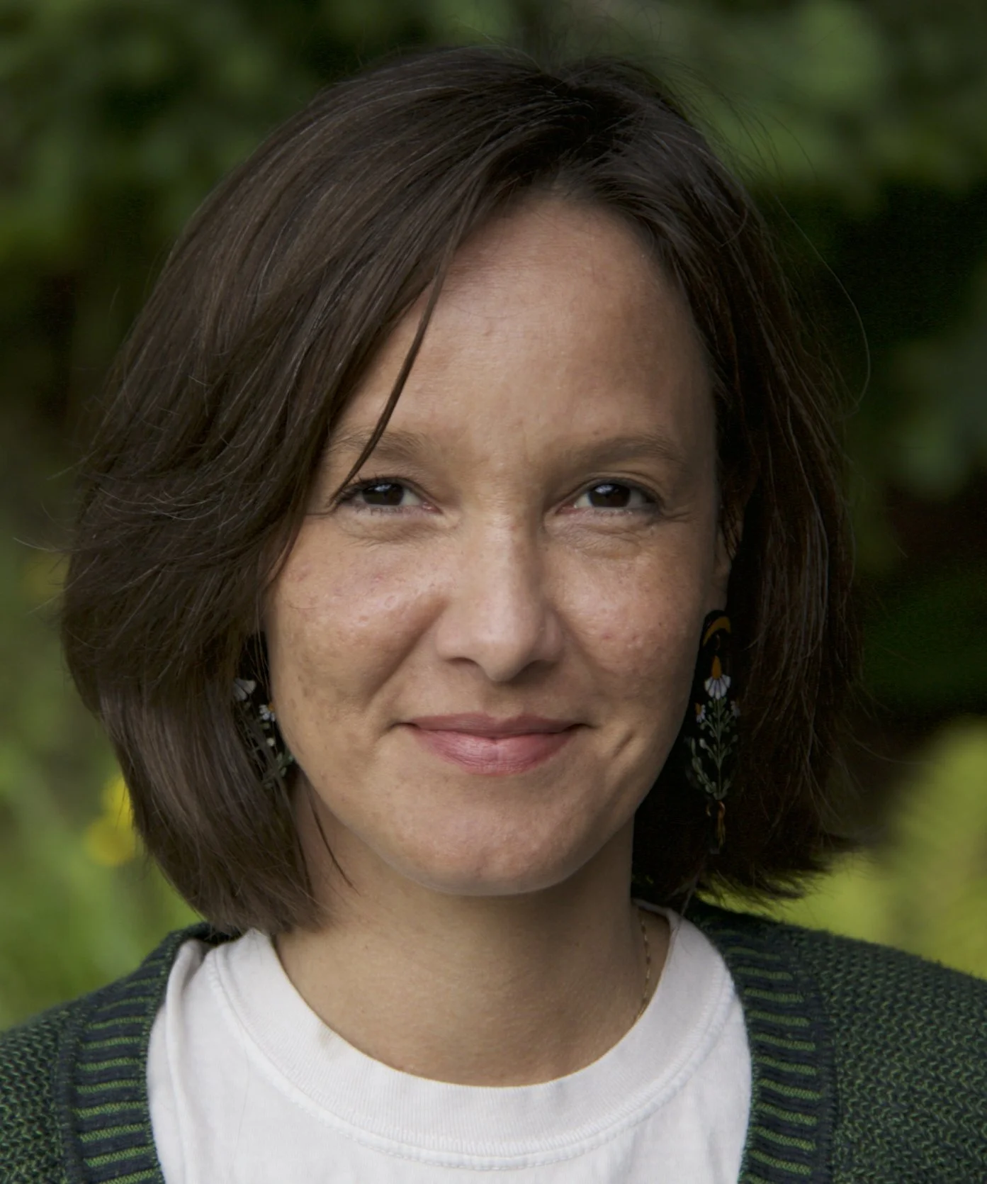 Close-up of a woman with shoulder-length brown hair, wearing earrings and a green sweater over a white shirt, standing outdoors with a blurred green background.