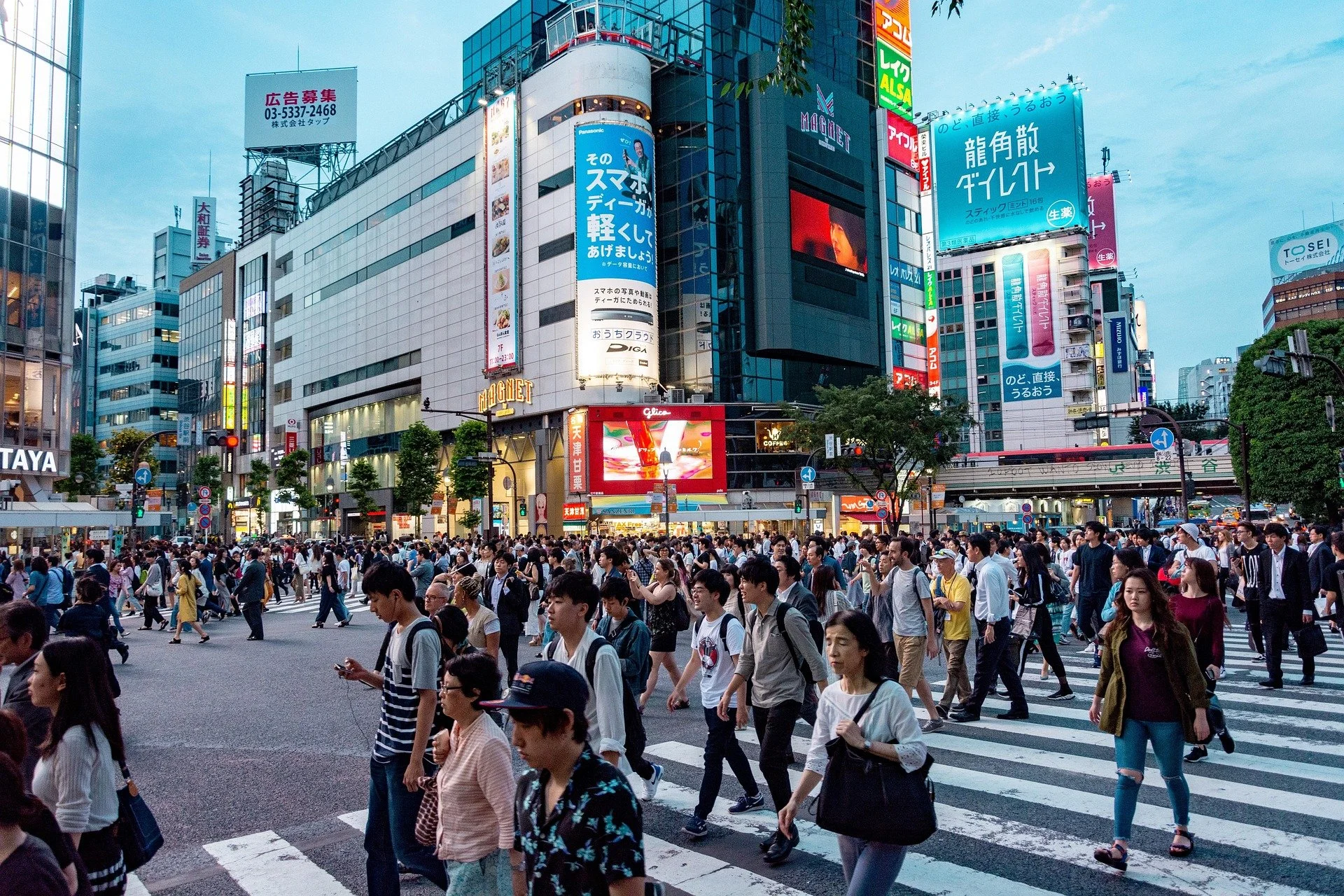 Crowd of people crossing the street in a busy urban area, surrounded by tall buildings and colorful advertising billboards in Tokyo.
