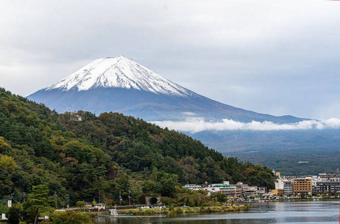 Snow-capped Mount Fuji in the background above a forested hillside and a small town by a body of water.