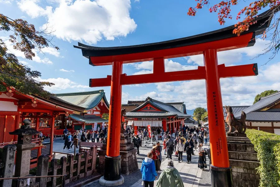 Crowd of people visiting a traditional Japanese shrine with a prominent red torii gate in the foreground, under a blue sky with white clouds.