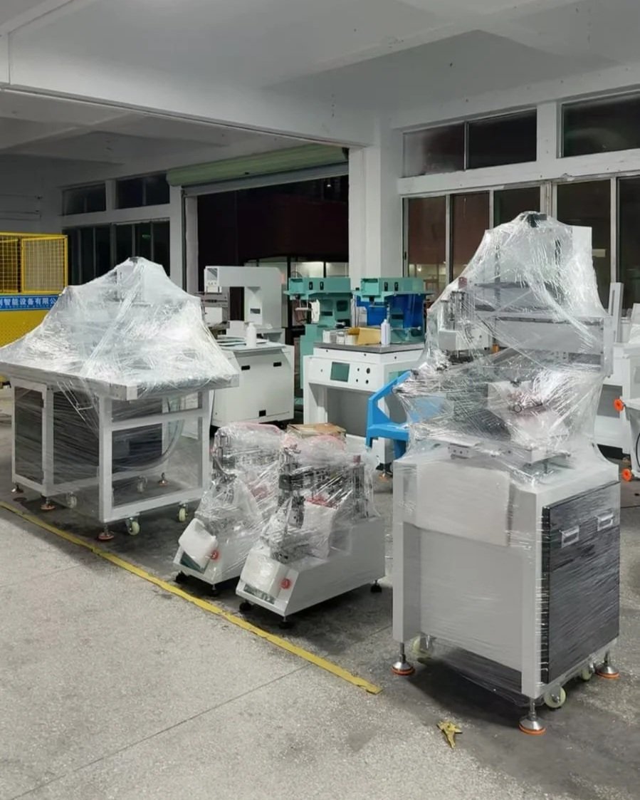 Several pieces of industrial equipment covered in plastic wrap are located inside a large room with windows. The equipment appears to be new or unused.