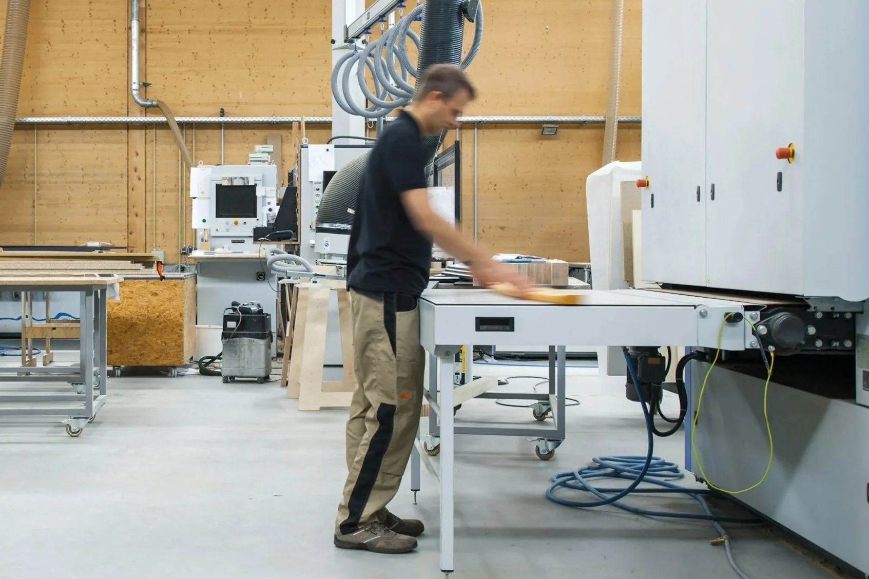 A man working in a woodworking shop, sanding a piece of wood on a conveyor belt among various woodworking machines and tools.