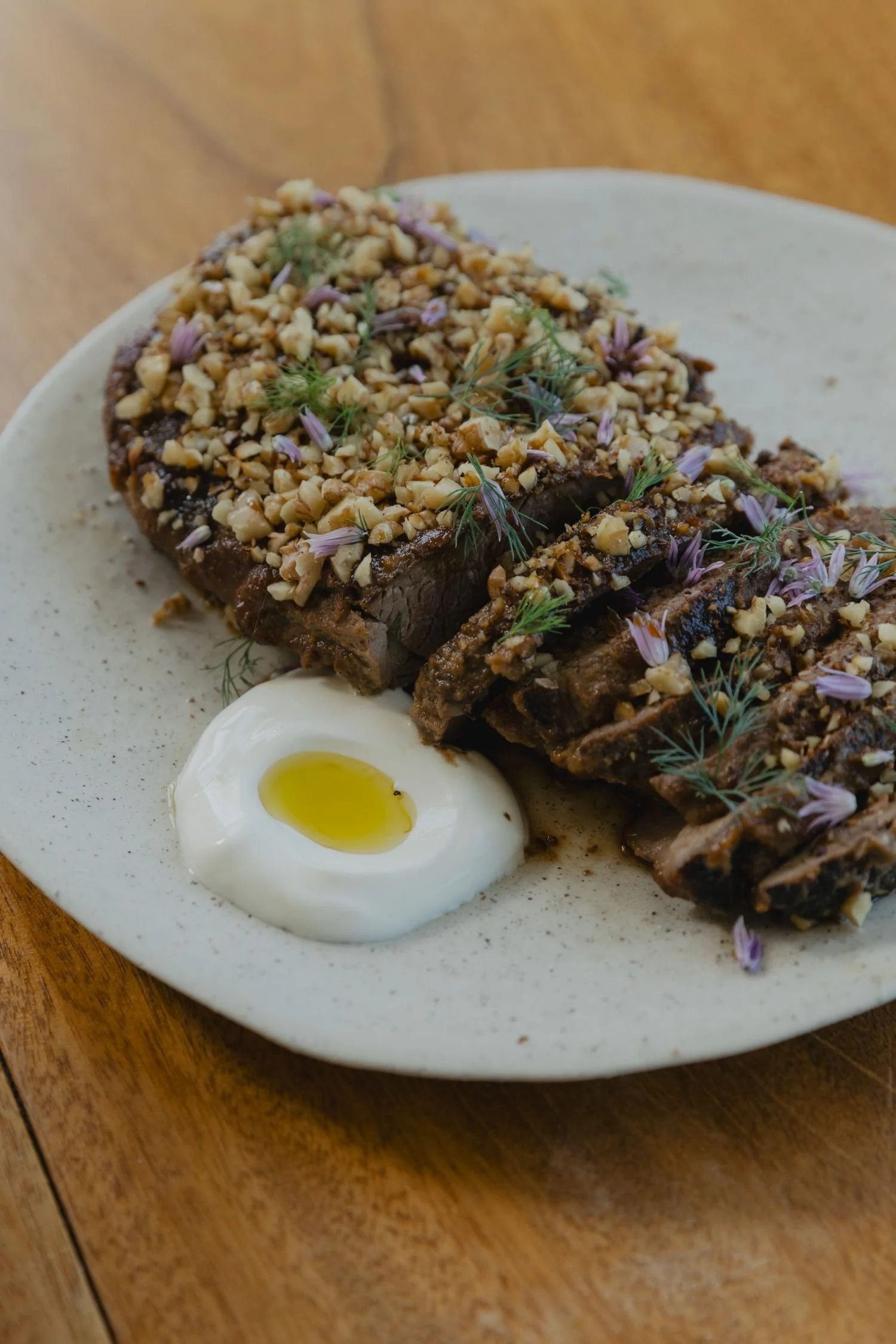 Slices of beef topped with crushed nuts and edible flowers, served with a soft-boiled egg with a runny yolk, on a white plate.