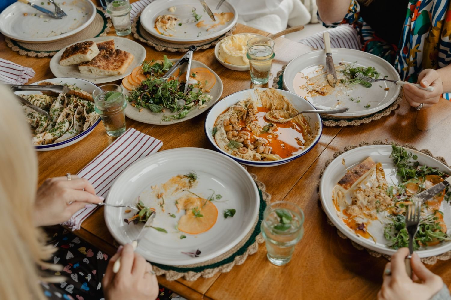 A table with mostly empty plates of remnants from a meal, mostly salad and leftover food, and several glasses of water. People are eating and cleaning up.