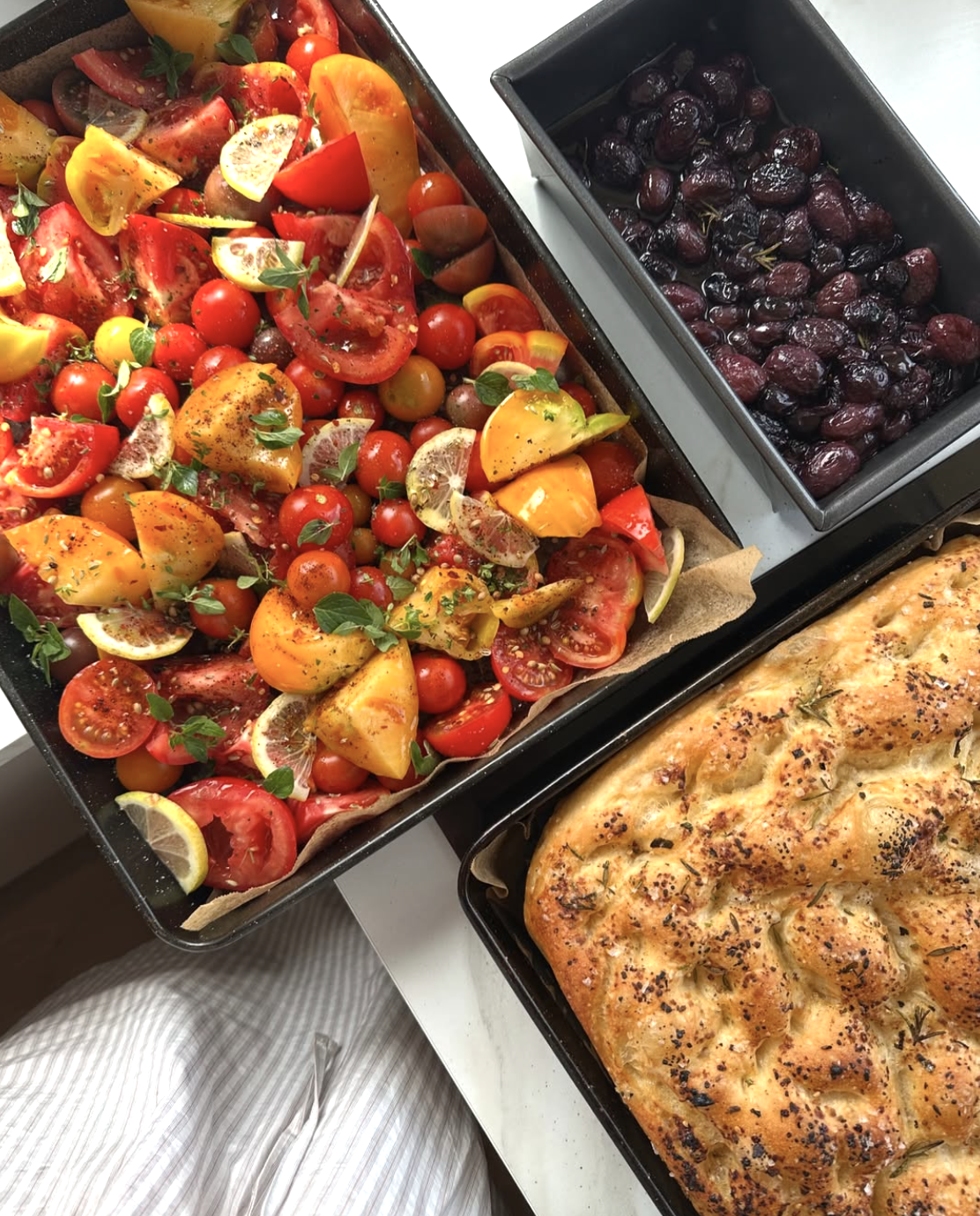 A tray of colorful tomato salad with lemon, basil, and herbs, a bowl of dark purple olives with sprigs of rosemary, and a roasted bread with herbs and seeds.