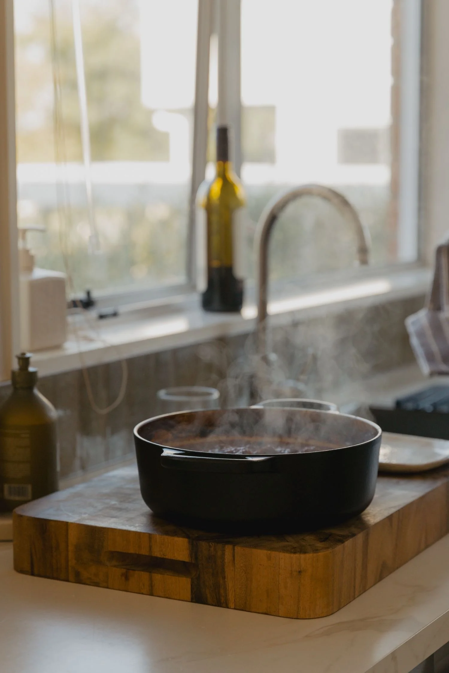 A black pot on a white kitchen countertop with a wooden base, in front of a window with a view outside, and a bottle of oil or vinegar, a soap dispenser, and a kitchen sink faucet in the background.