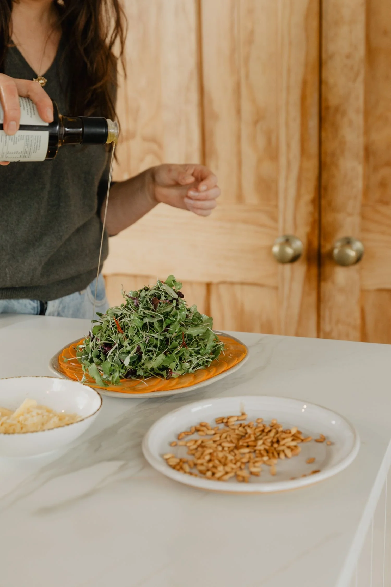 A person pouring dressing or oil onto a salad with greens on a decorative orange plate on a white table. There are additional small bowls with other ingredients, such as shredded cheese and sunflower seeds. The background features wooden cabinets.