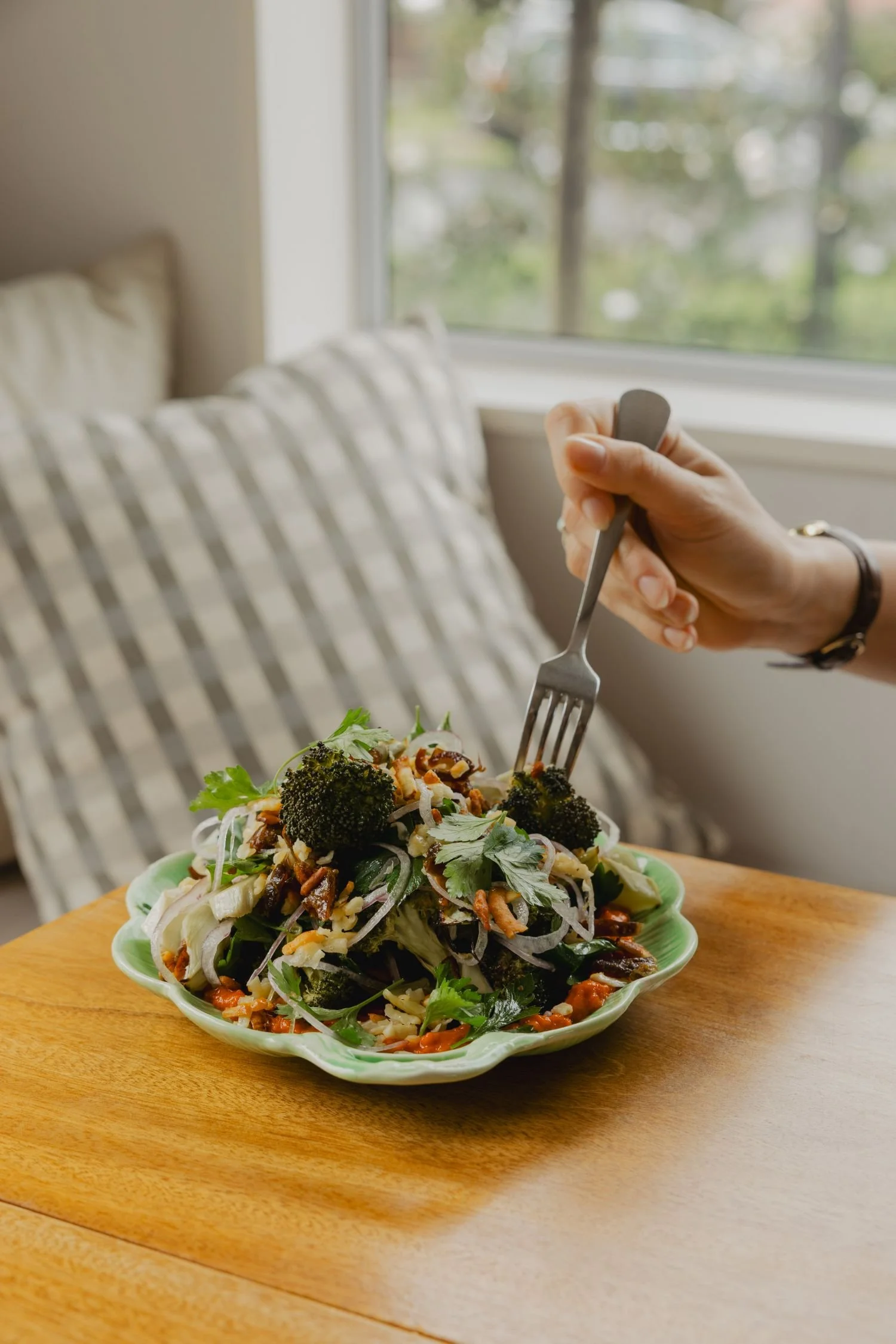 Hand holding a fork over a plate of vegetable salad on a wooden table, with a window and striped curtain in the background.