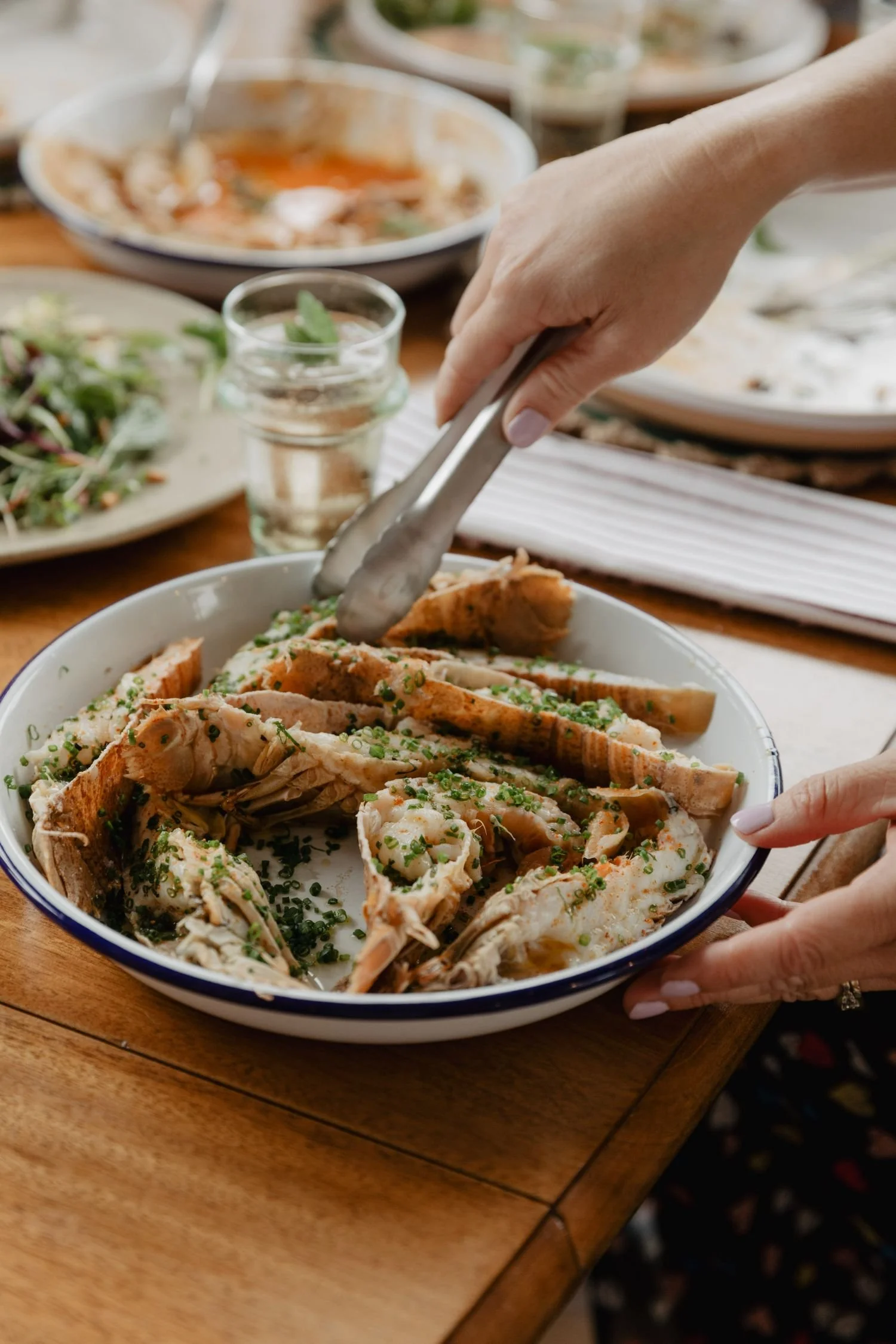 A person serving cooked fish garnished with chopped herbs on a white plate with a blue rim at a wooden table, with various dishes and a glass of water in the background.