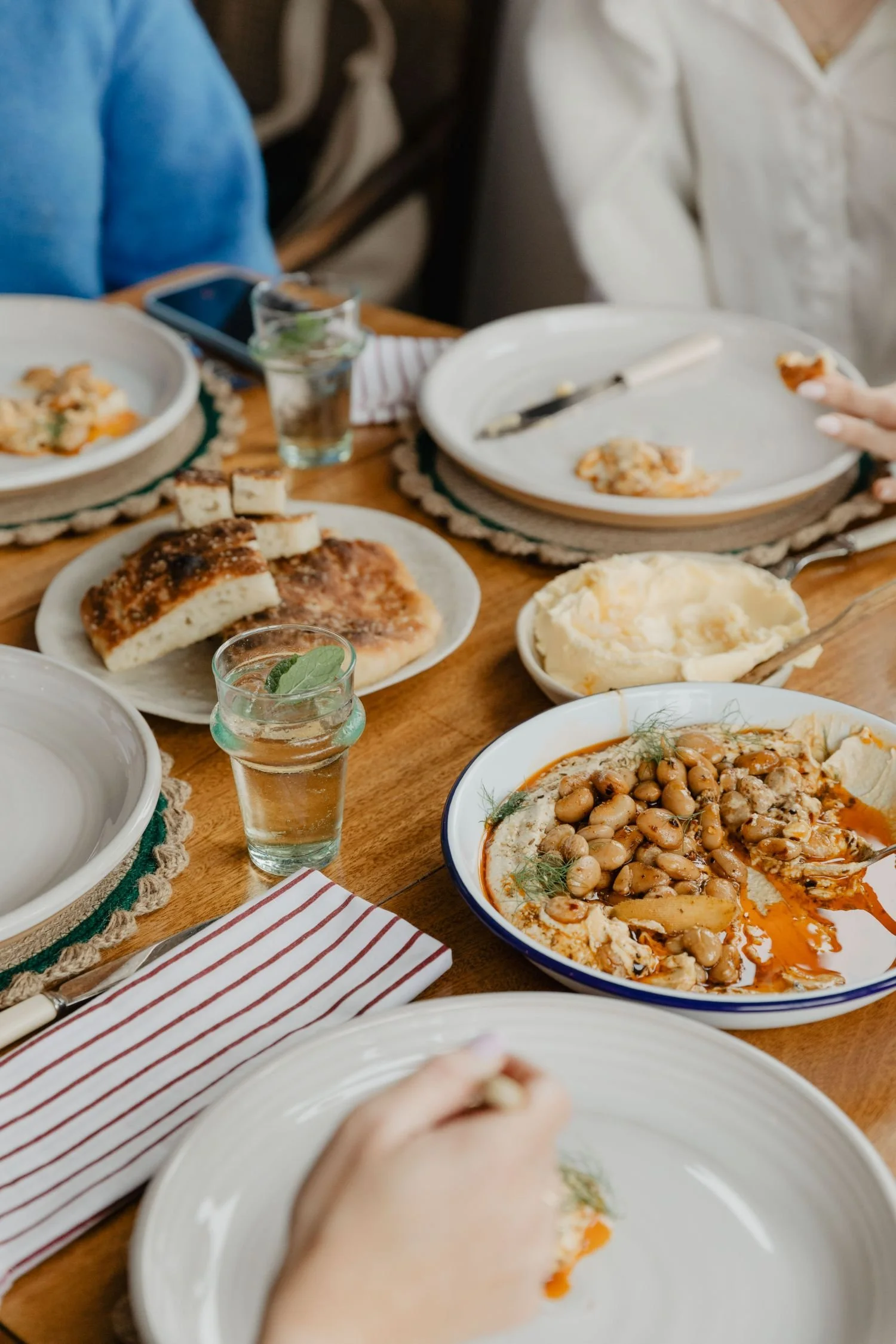 A close-up of a dinner table set with various plates of food, including baked bread, a bean dish, and other meals. There are glasses of water, utensils, a striped napkin, and partially visible plates and hands of people dining.