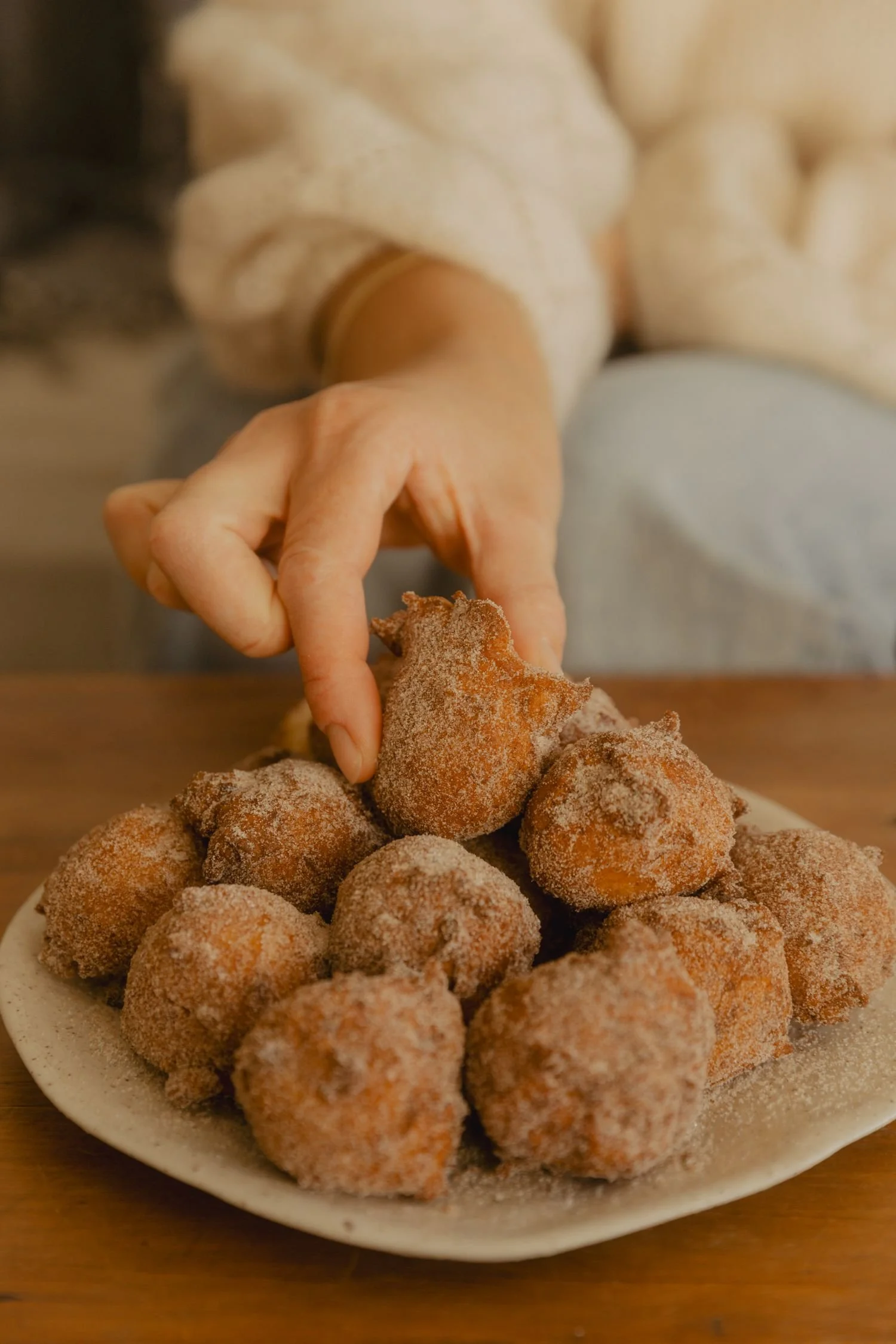 A white plate filled with coated fried croquettes or similar round snacks dusted with powder, likely sugar or cinnamon, on a wooden surface.