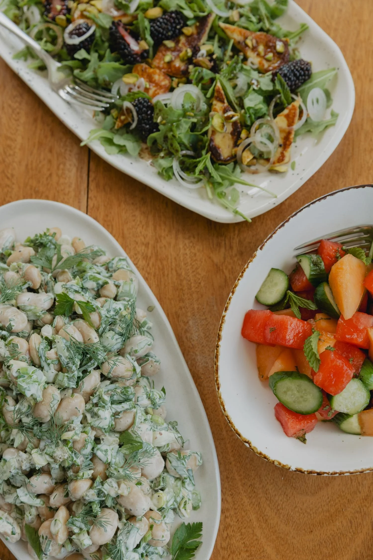 A platter of chickpea salad garnished with herbs and a bowl of watermelon, cucumber, and tomato salad on a wooden table.