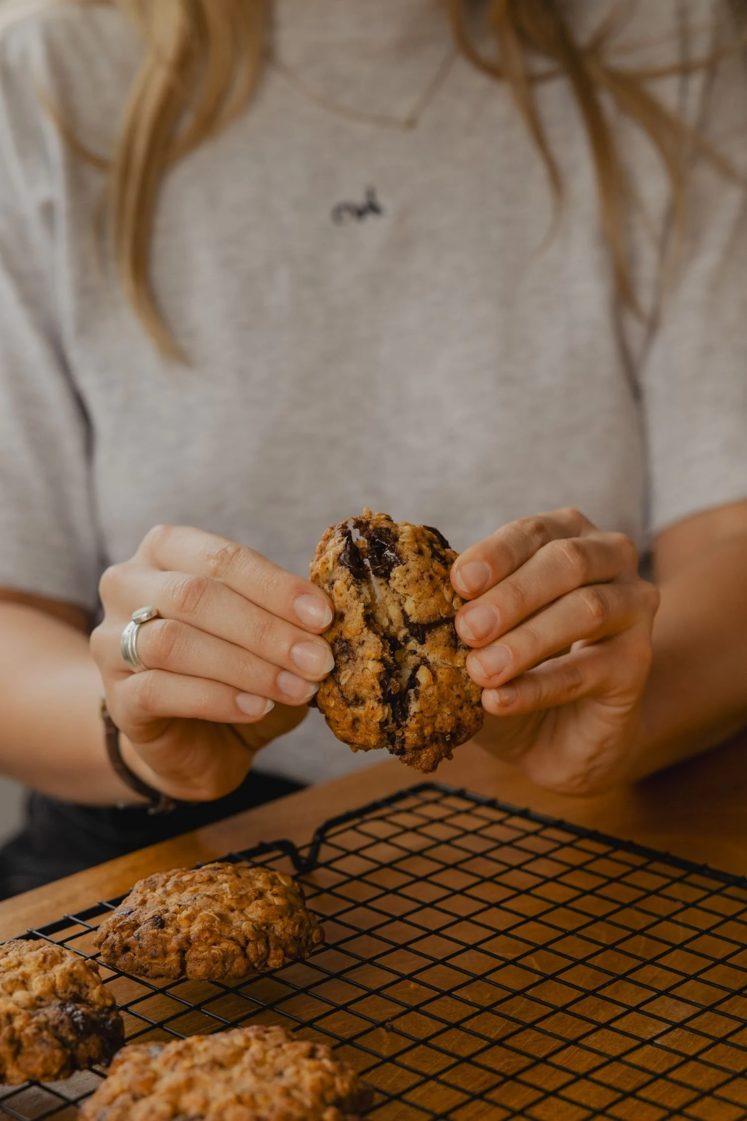 Person holding a large chocolate chip cookie, with more cookies on a cooling rack on a wooden table.