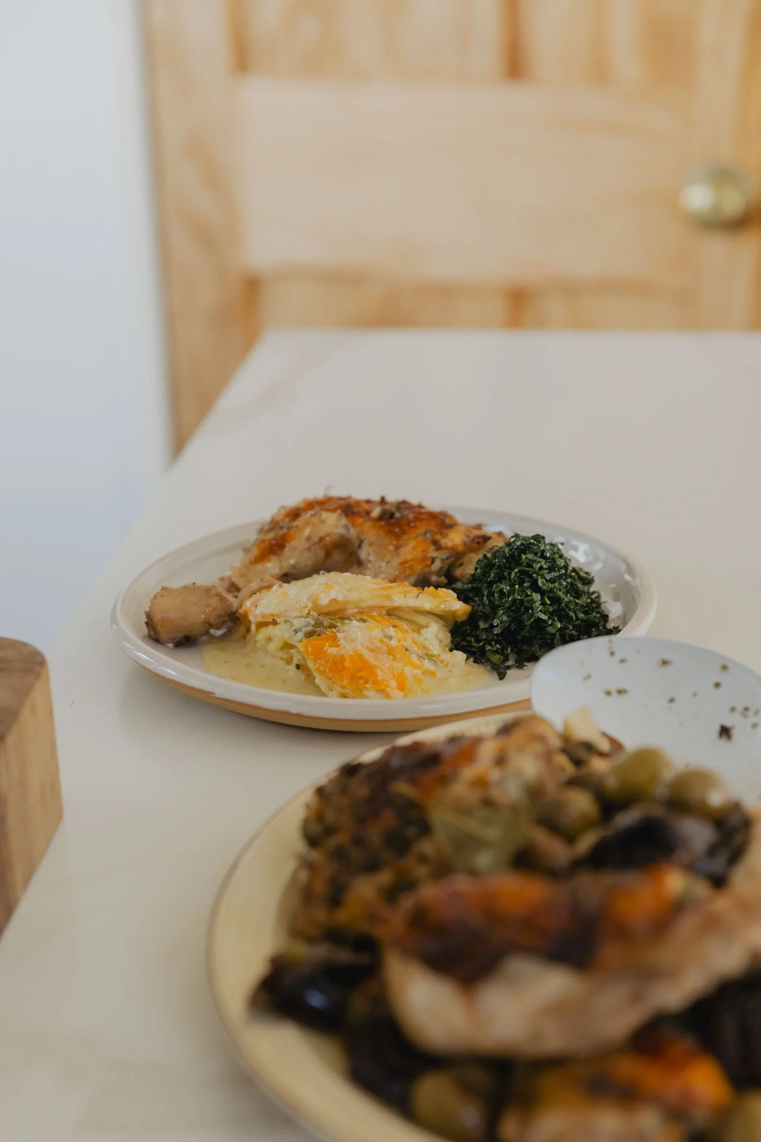 A white plate with eggs, chicken, black leafy greens, and potatoes on a white table, with a wooden chair in the background.