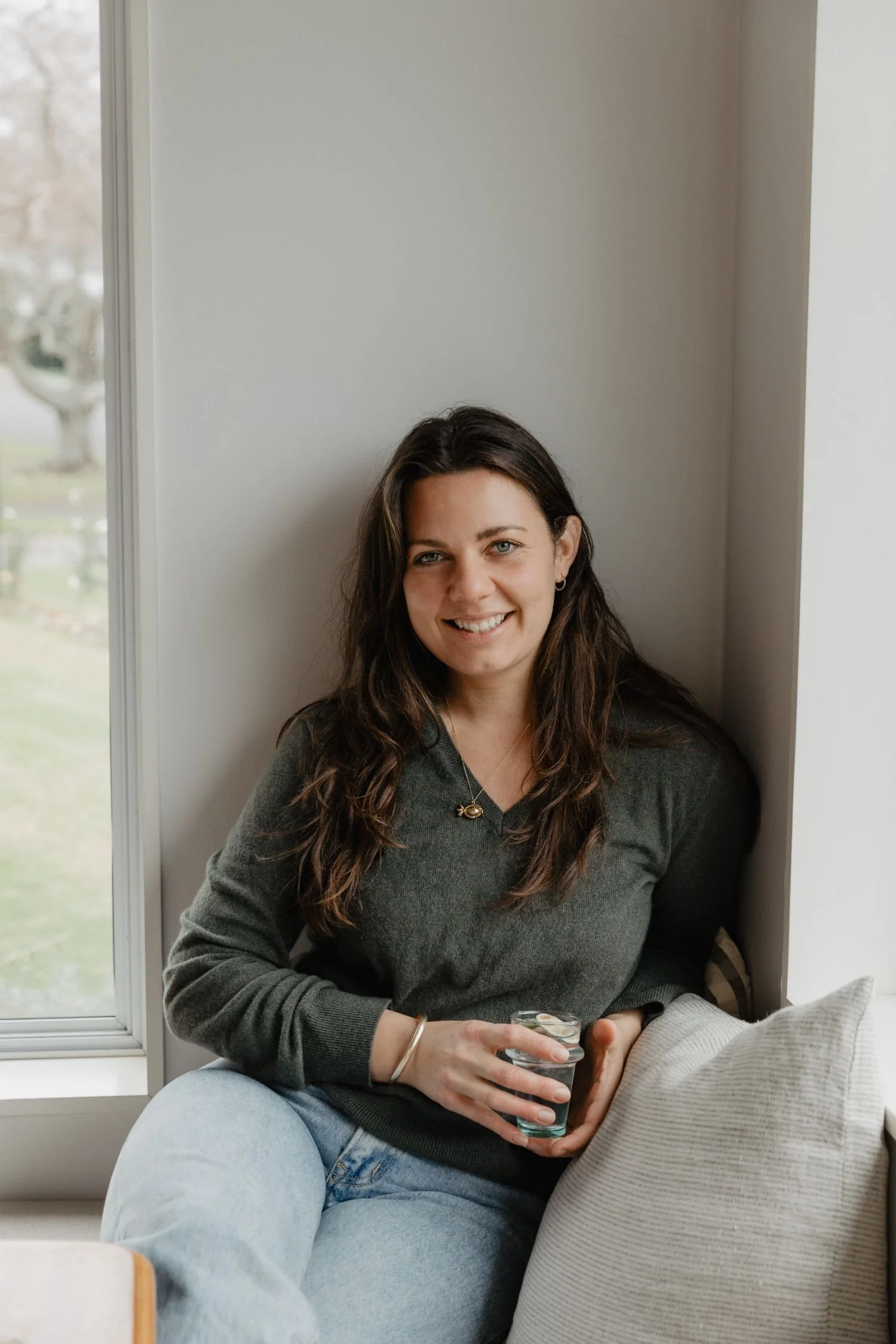 A woman with long dark hair and blue eyes smiling at the camera, sitting by a window, holding a glass of water, wearing a dark grey sweater and jewelry, in a cozy indoor setting.