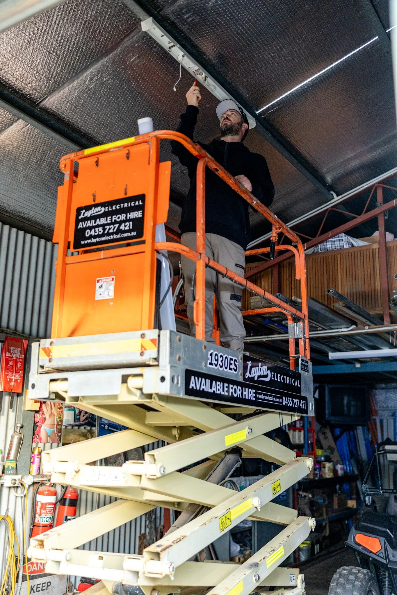 A man working on electrical wiring on a ceiling while standing on an orange scissor lift in a hardware store. The lift has a black sign indicating it is available for hire, with contact information. The background shows store shelves and a fire extinguisher.