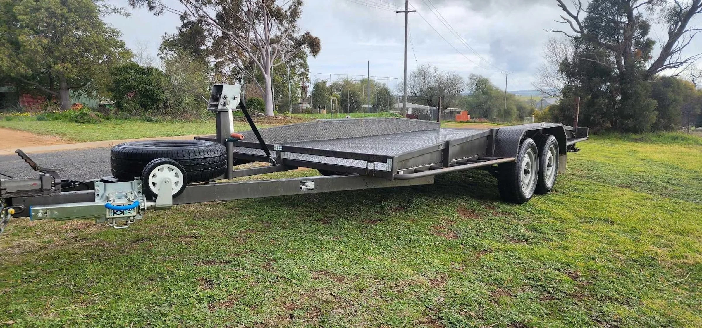 A black utility trailer with dual wheels, a spare tire, and a metal platform, parked on grass near a suburban street with trees and power lines.