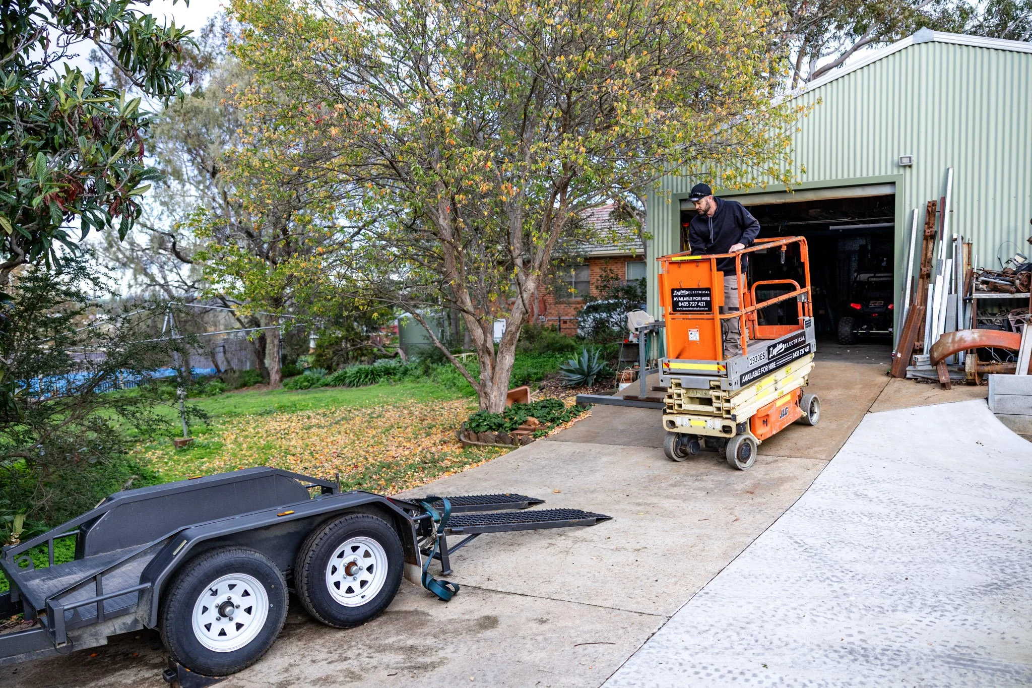A man operating a small orange lift in a backyard next to a green shed, with trees and a grassy area in the background.
