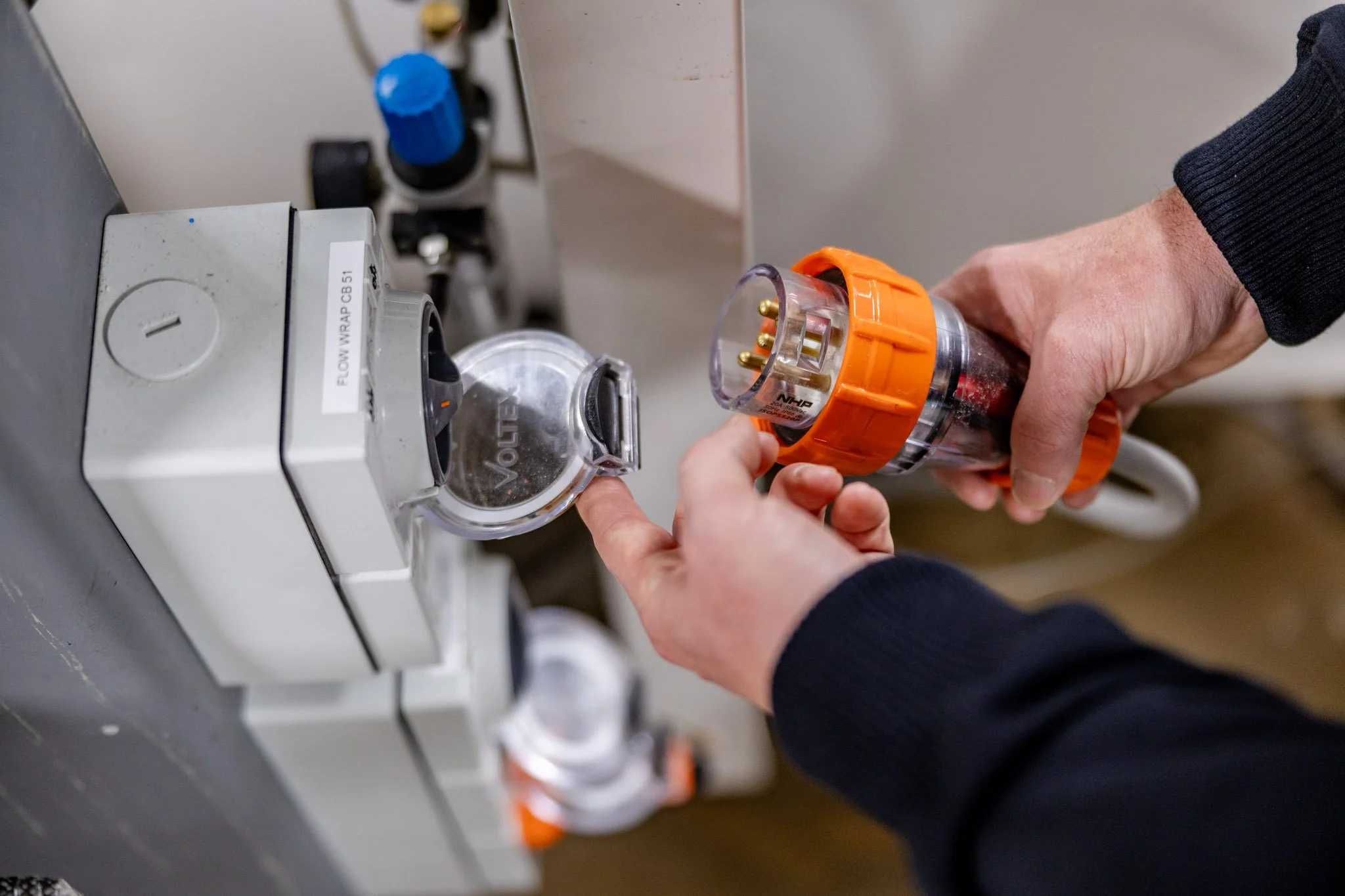 A person is holding a clear plastic container with orange and black accents, containing electrical pins, and is inserting it into a socket on a piece of equipment.
