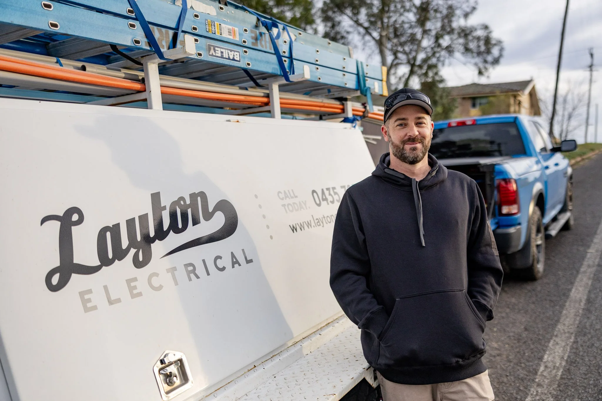 Mitch standing outdoors next to a white electrical service truck with ladders mounted on top and a large sign that reads 'Layton Electrical'.  In the background, there is a blue pickup truck and some trees, with a partly cloudy sky.