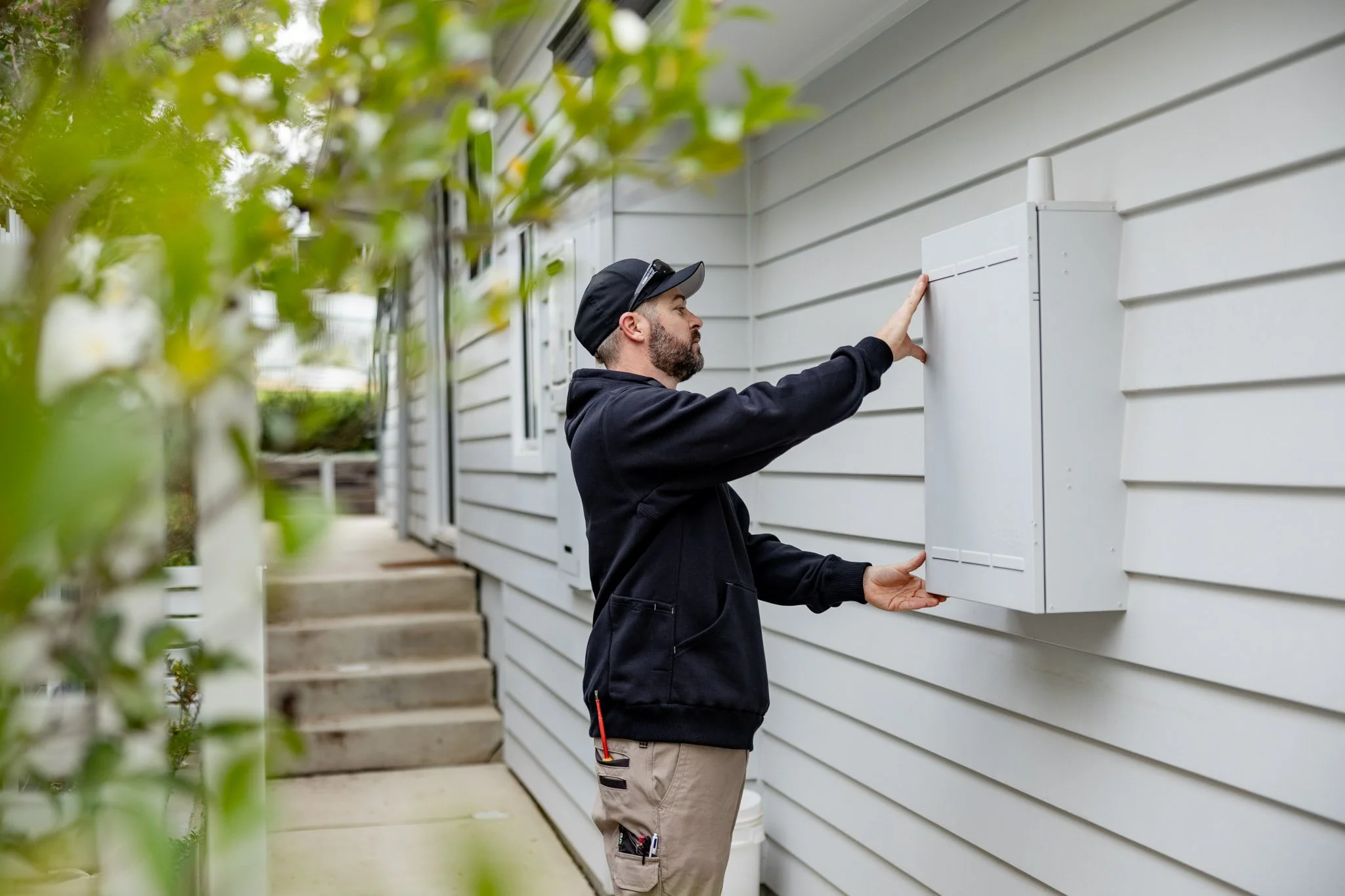 A man wearing a black hoodie, beige pants with tools attached, black cap, and sunglasses on his head installing or inspecting a white electrical or communication box outside a house with beige horizontal siding. Green foliage partially obscures the view in the foreground.