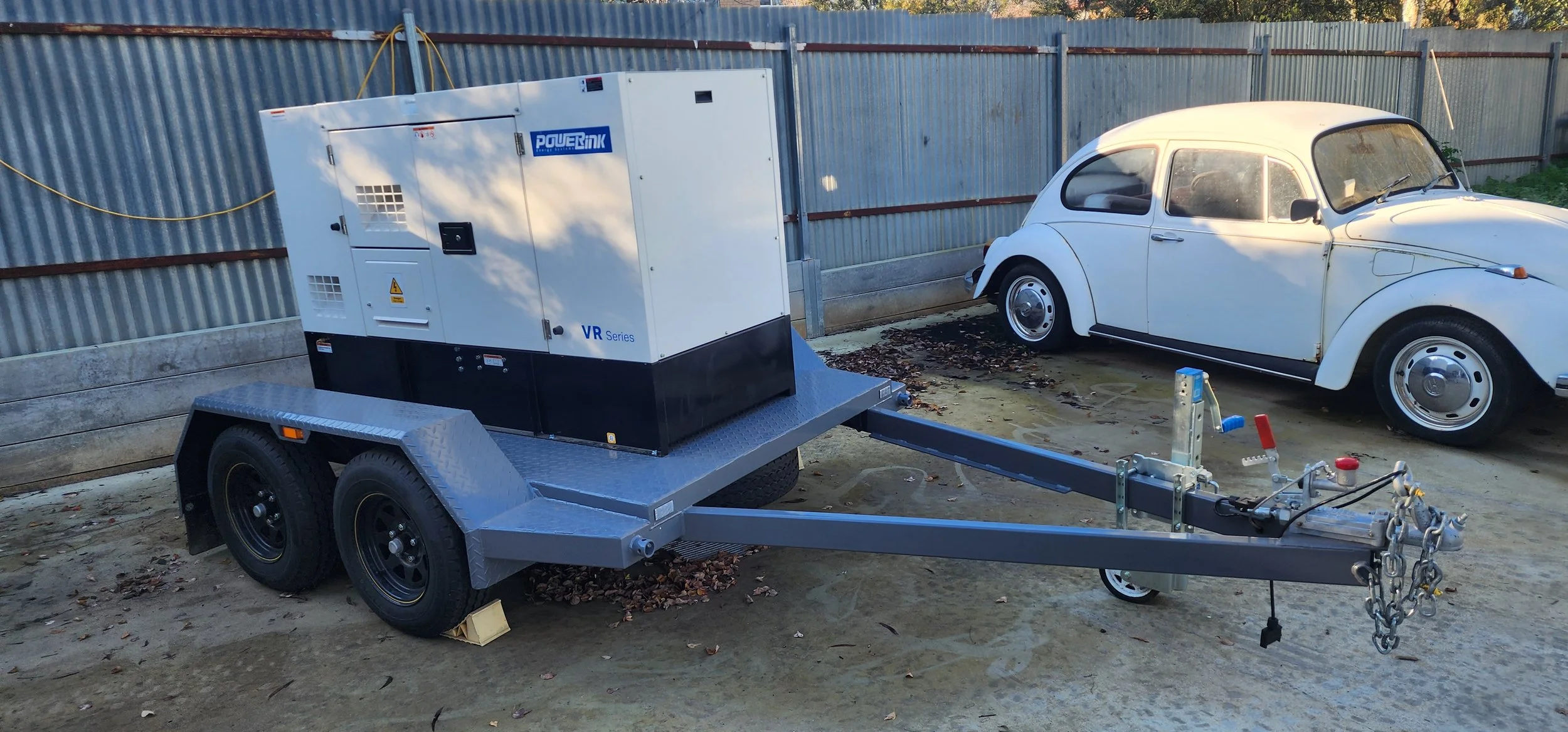 A white vintage Volkswagen Beetle car parked behind a large white portable generator on a trailer in an outdoor area with a corrugated metal fence and fallen leaves on the ground.