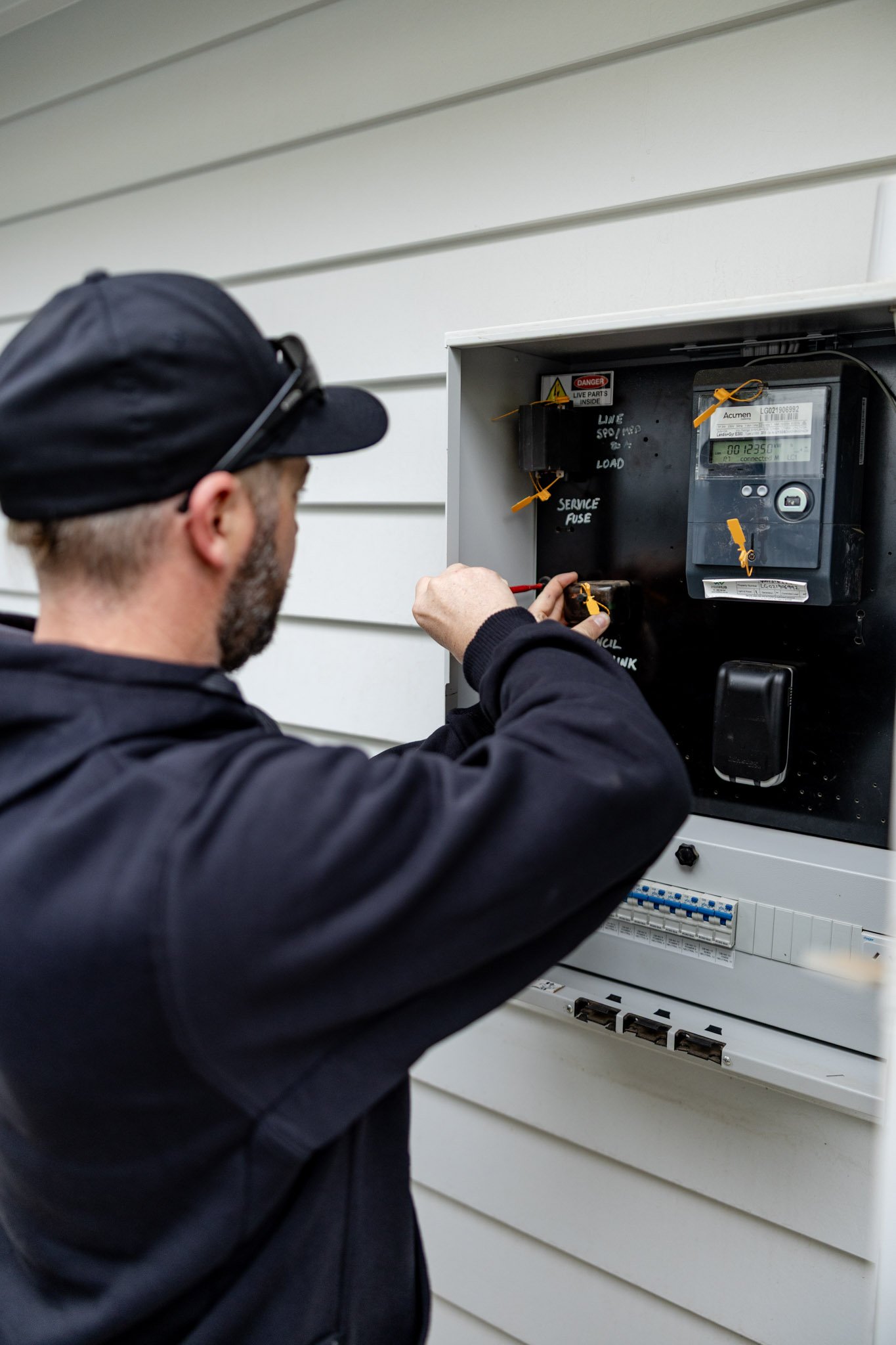 A technician wearing a black cap and jacket working on an electrical panel, connecting wires with a screwdriver outside a house with white siding.