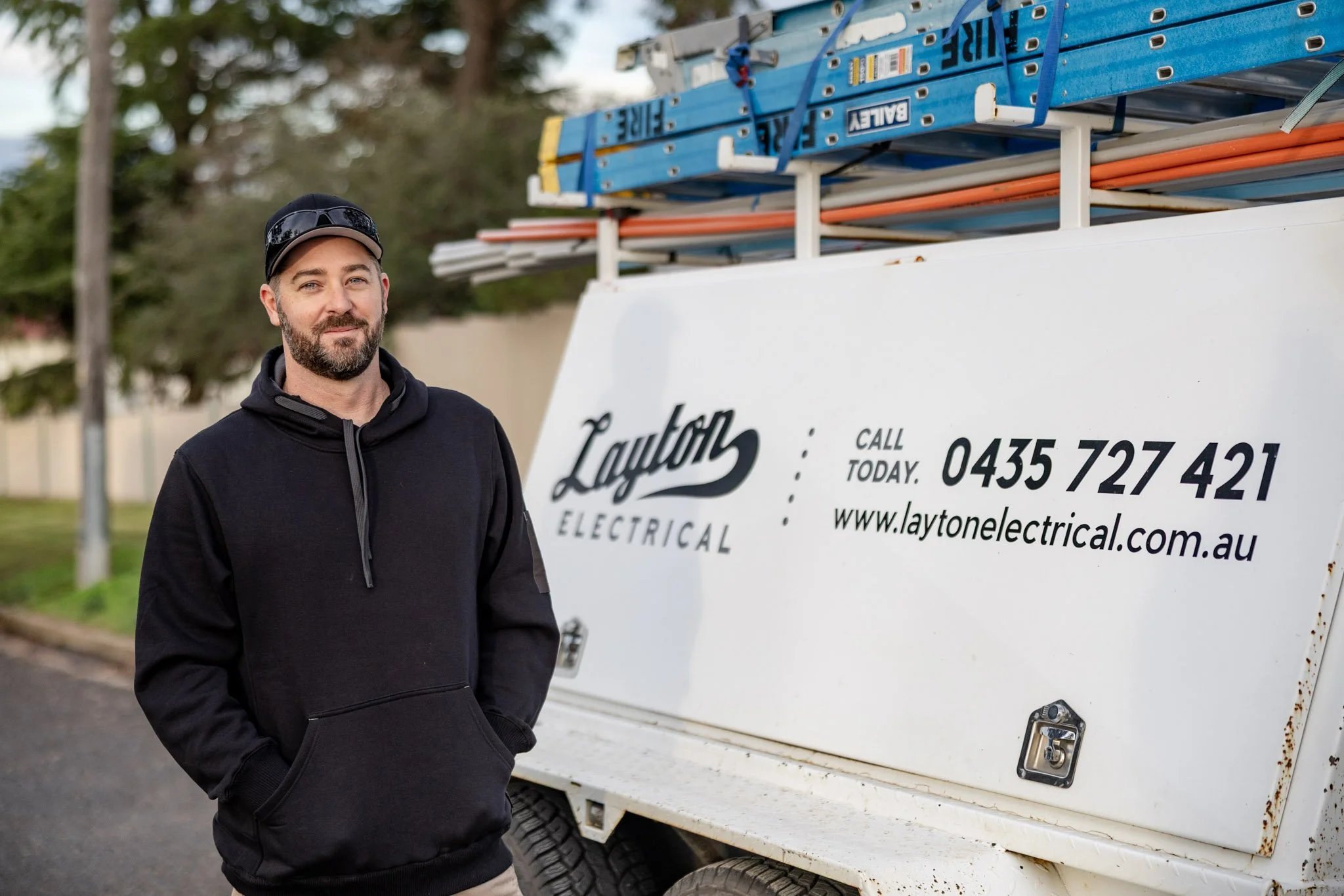 Mitch standing next to a white utility vehicle with ladder and electrical tools on top, advertising Layton Electrical, with trees in the background.