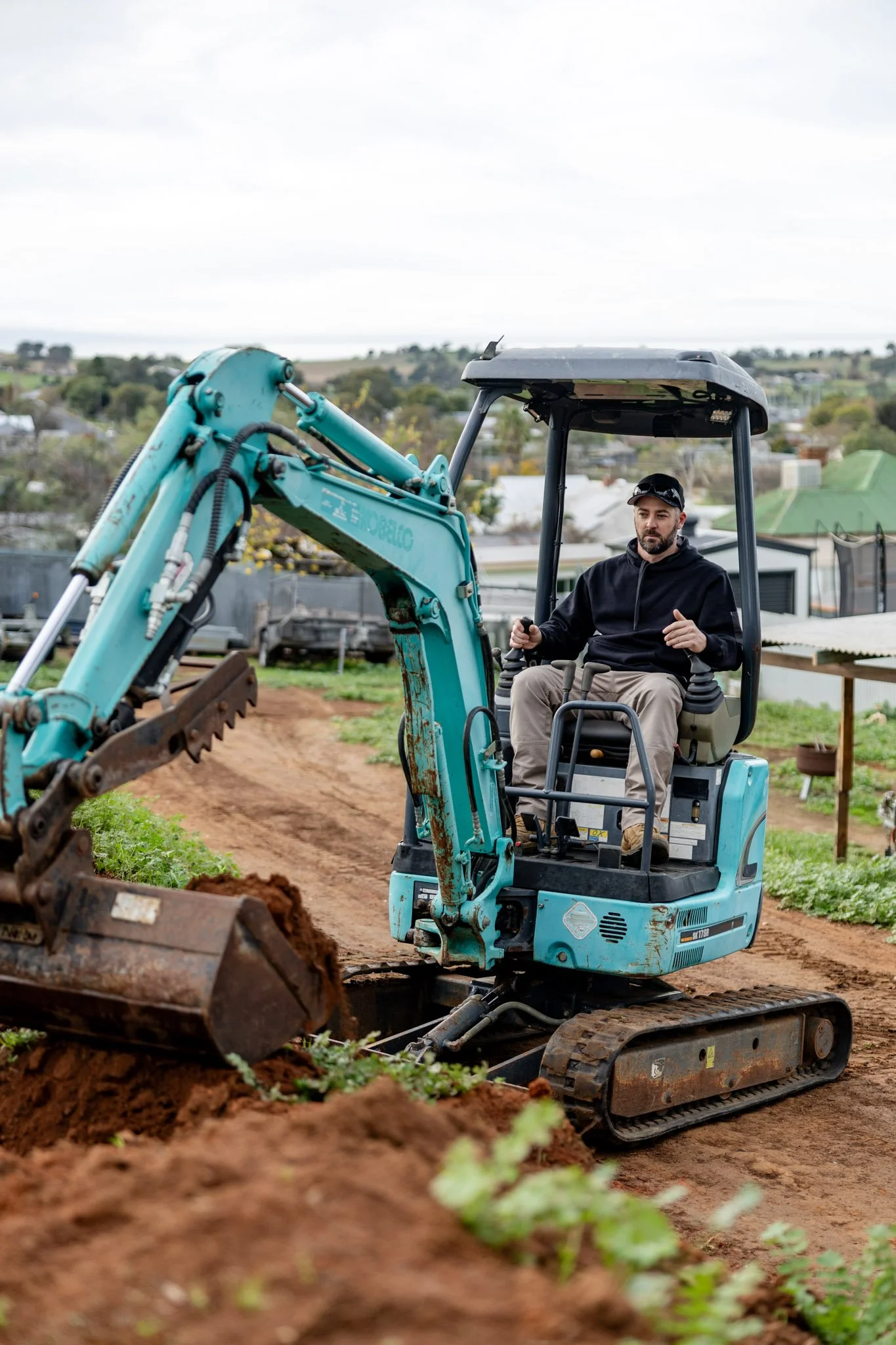 A man operating a small blue excavator on a farm or garden, with trees and buildings in the background, overcast sky.