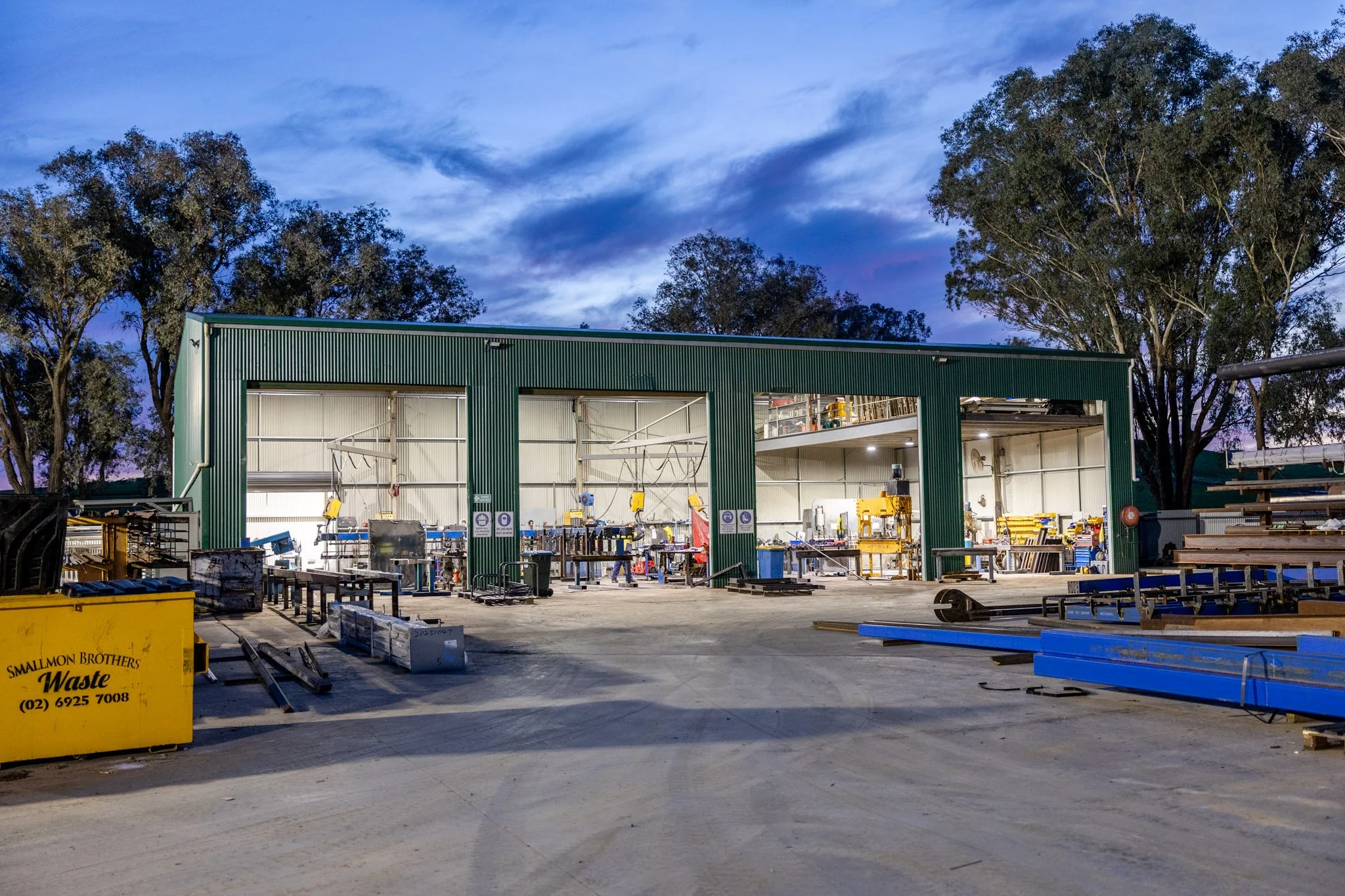 An industrial workshop with a green metal building, tools, and equipment inside and outside, surrounded by trees under a partly cloudy evening sky.