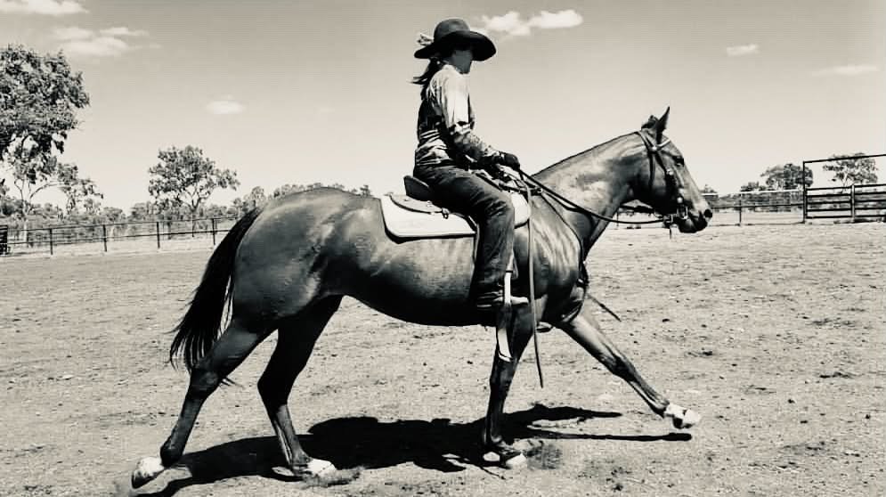A woman riding a horse in a saddle on a dirt field, with trees and a fence in the background, in black and white.