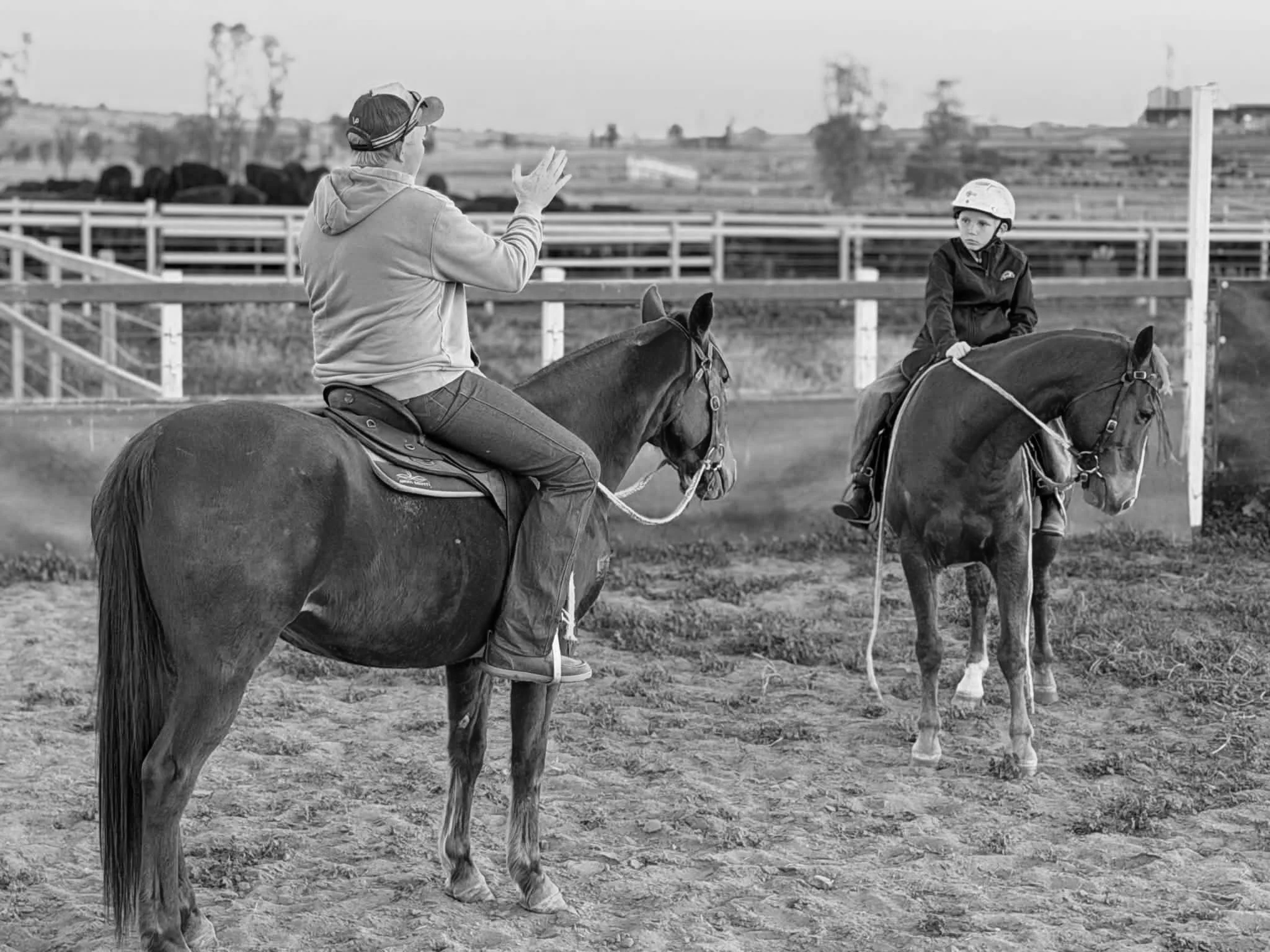 A man and a child riding horses on a dirt field in an outdoor equestrian area, with fencing and trees in the background in black and white.