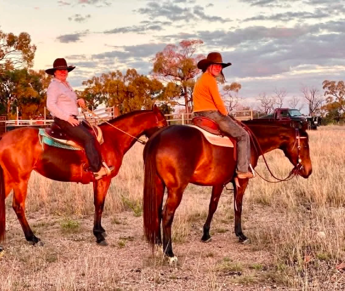 Two women riding brown horses in a field during sunset, wearing cowboy hats and casual clothing, with trees and a truck in the background.