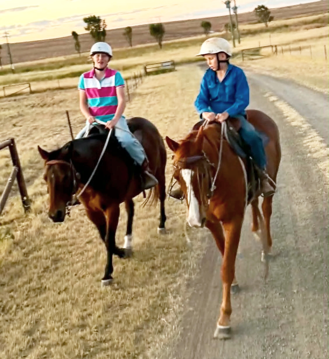 Two children riding horses on a dirt path in a rural area during sunset with open fields and trees in the background.
