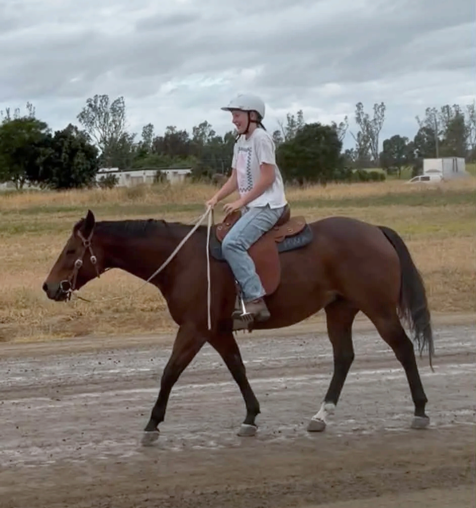 A person wearing a helmet riding a brown horse on a dirt path in a rural area with trees and overcast sky in the background.