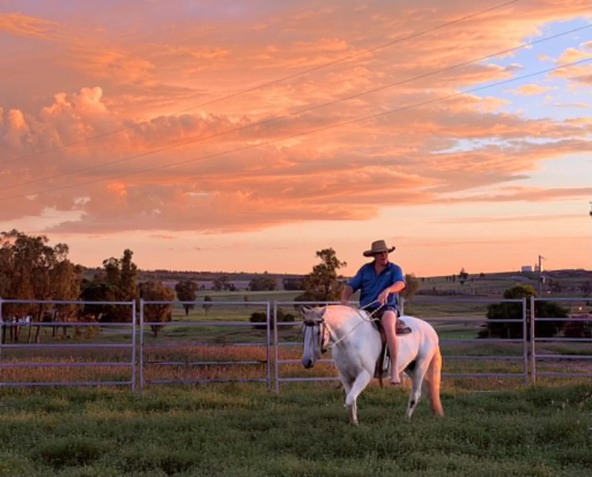 A person riding a white horse inside a fenced area during sunset with a colorful sky and scattered clouds.